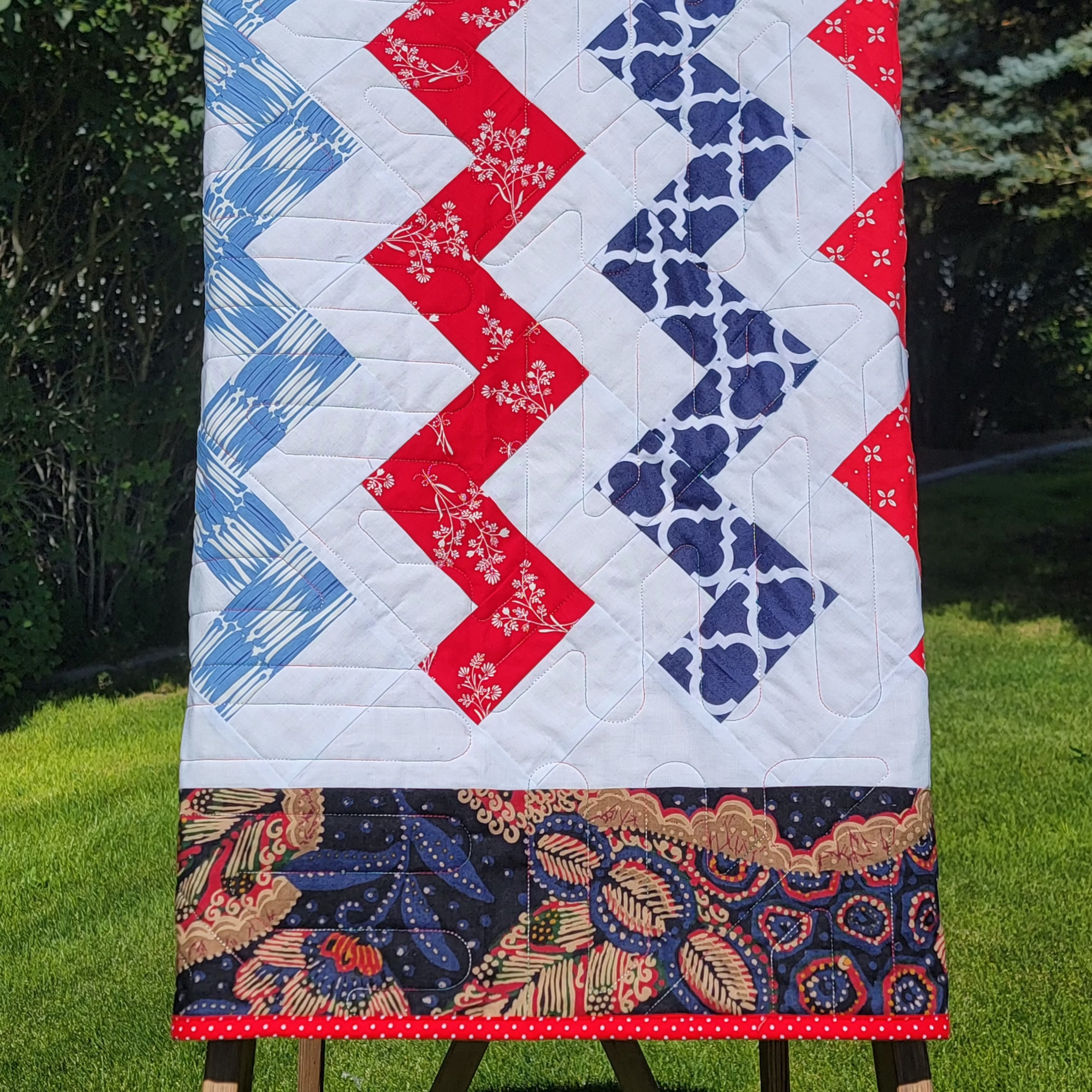 A handmade quilted baby blanket with a red, white, and blue chevron pattern and a floral border, draped over a wooden step ladder in a grassy backyard.