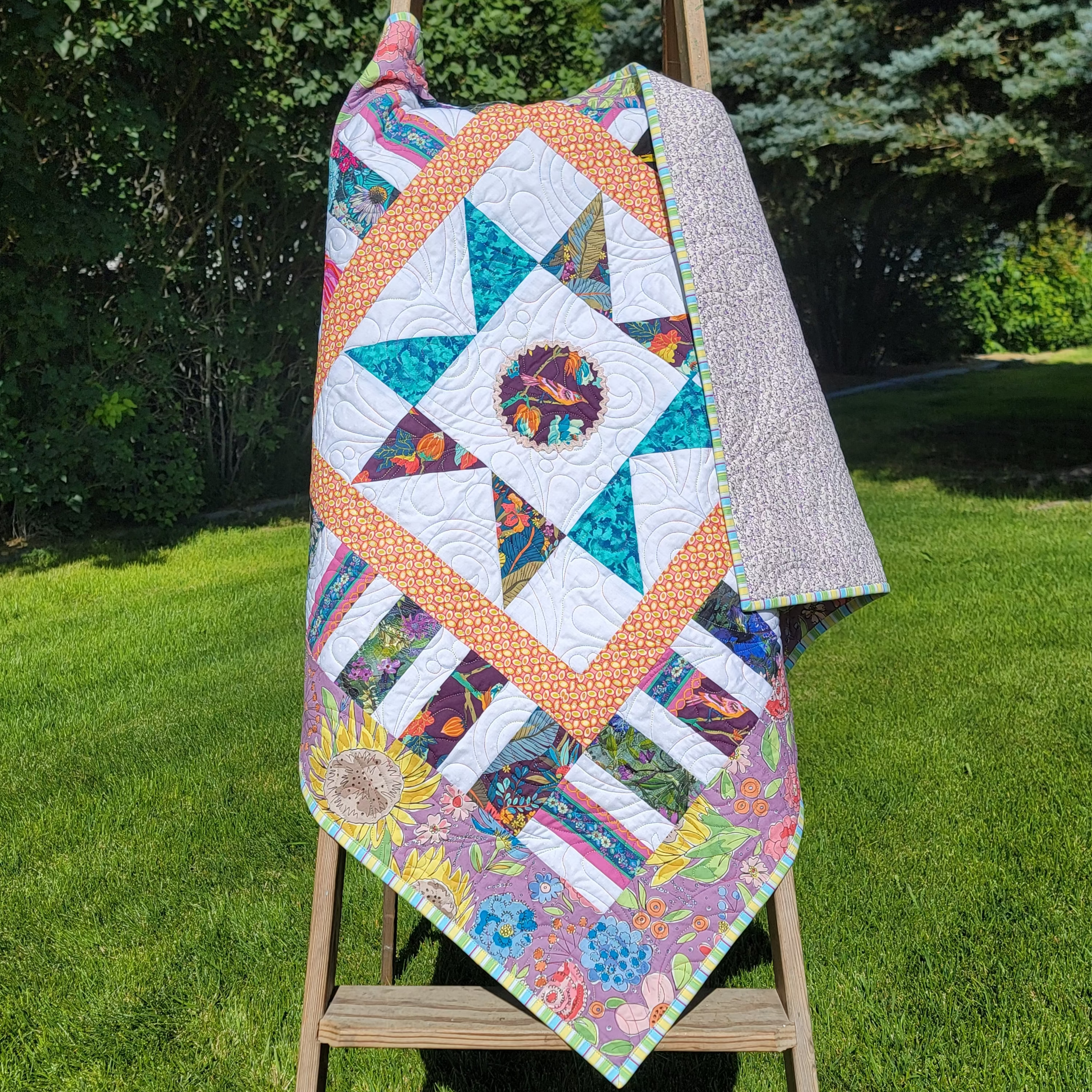 A colorful patchwork quilt draped over a wooden easel outdoors on a grassy lawn with trees in the background.