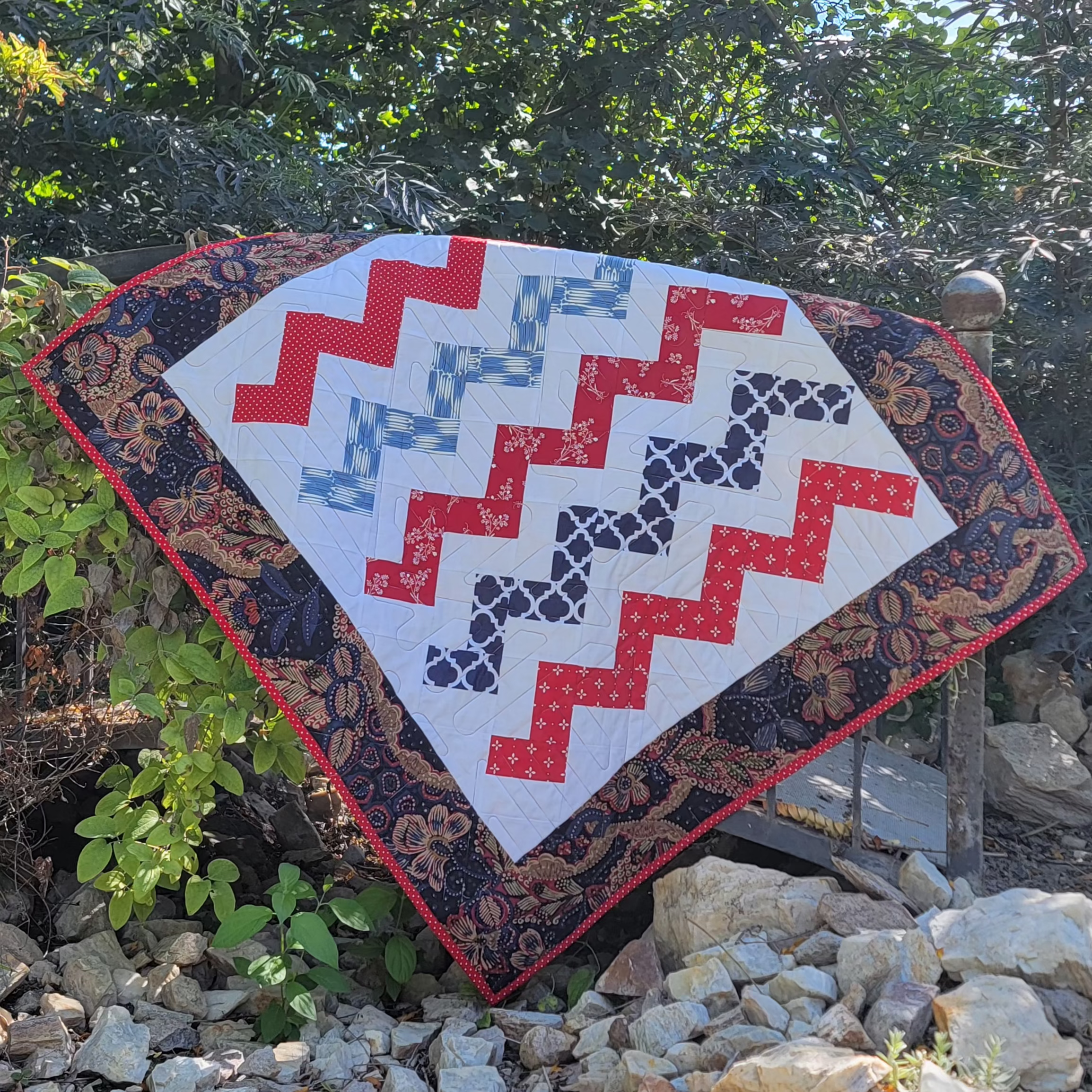 A handmade quilted patchwork blanket with a zigzag pattern in red, blue, and navy on a white background, surrounded by a dark floral border, displayed outdoors on a wooden stand among rocks and greenery.