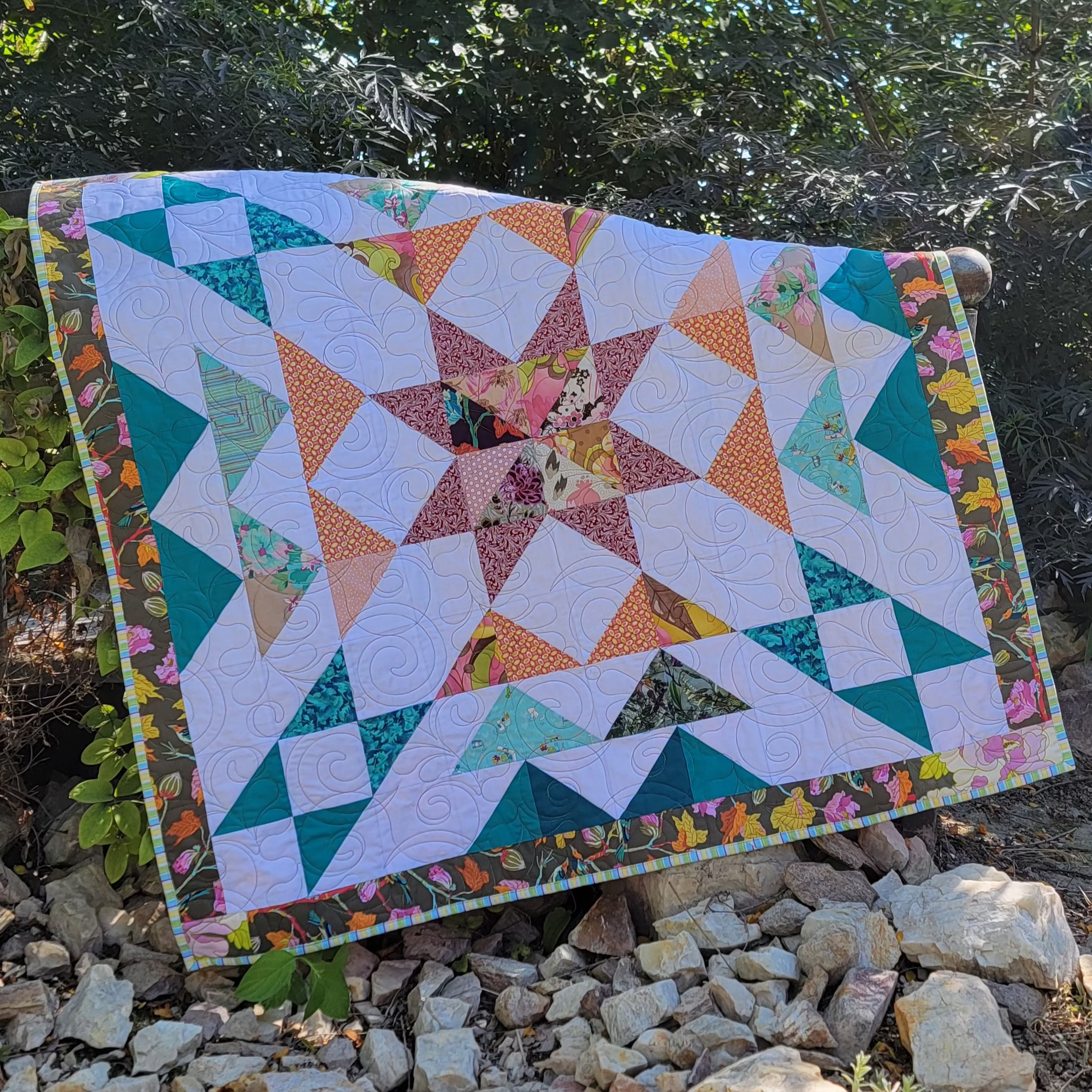A handmade quilt in white, teal, orange and burgundy colours in multiple prints with a central star draped over a wooden bridge in an outdoor setting. 