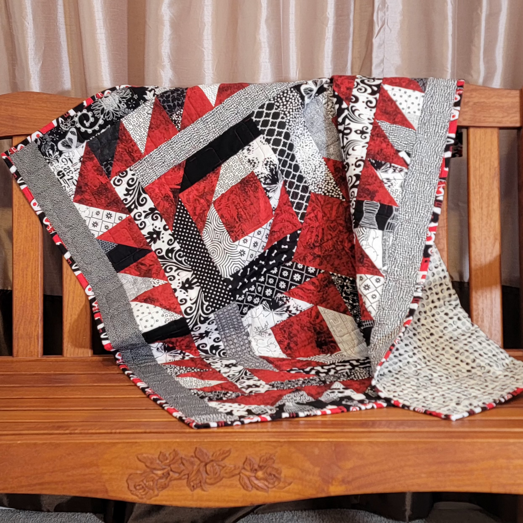 A red, white and black quilt draped over the back of a wooden bench.