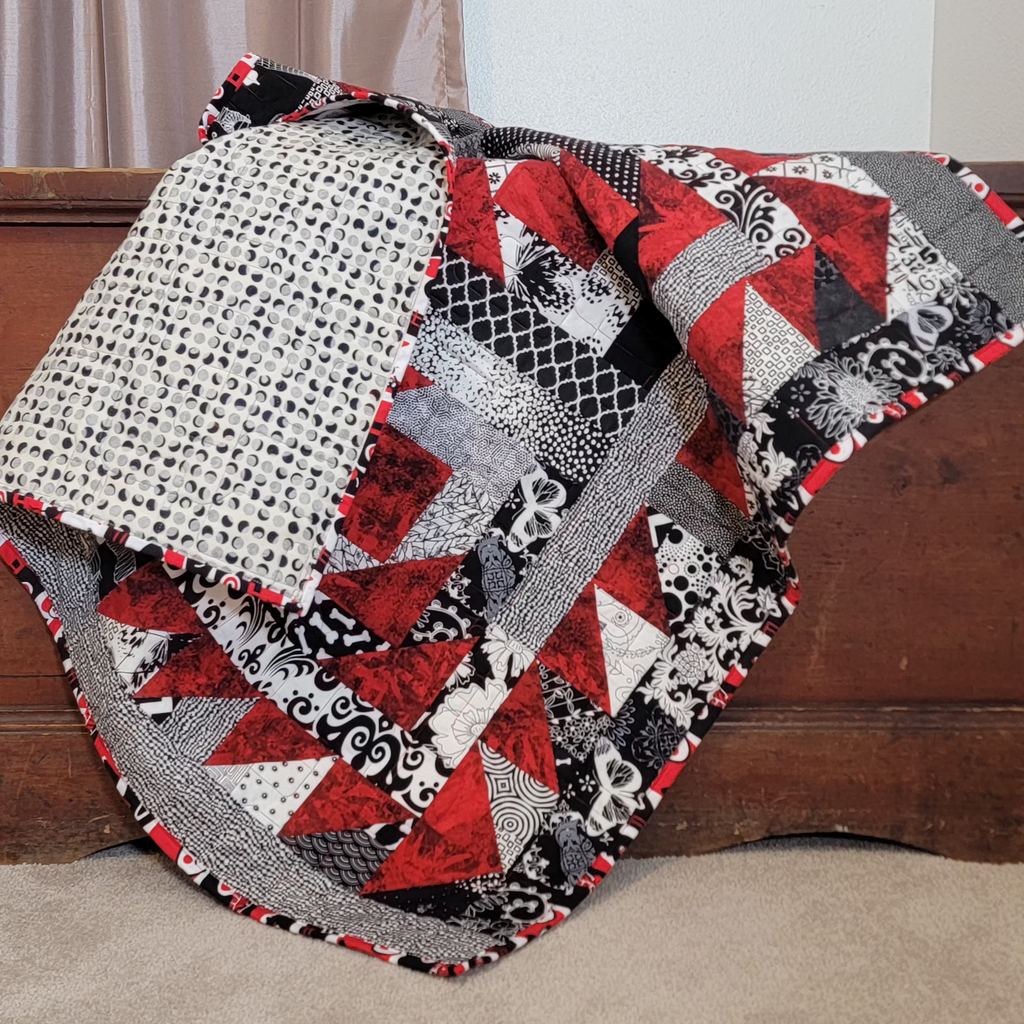 A red, white and black geometric quilt draped over a brown wooden chest, with a glimpse of the reverse side of the quilt in a black and white moon print. 