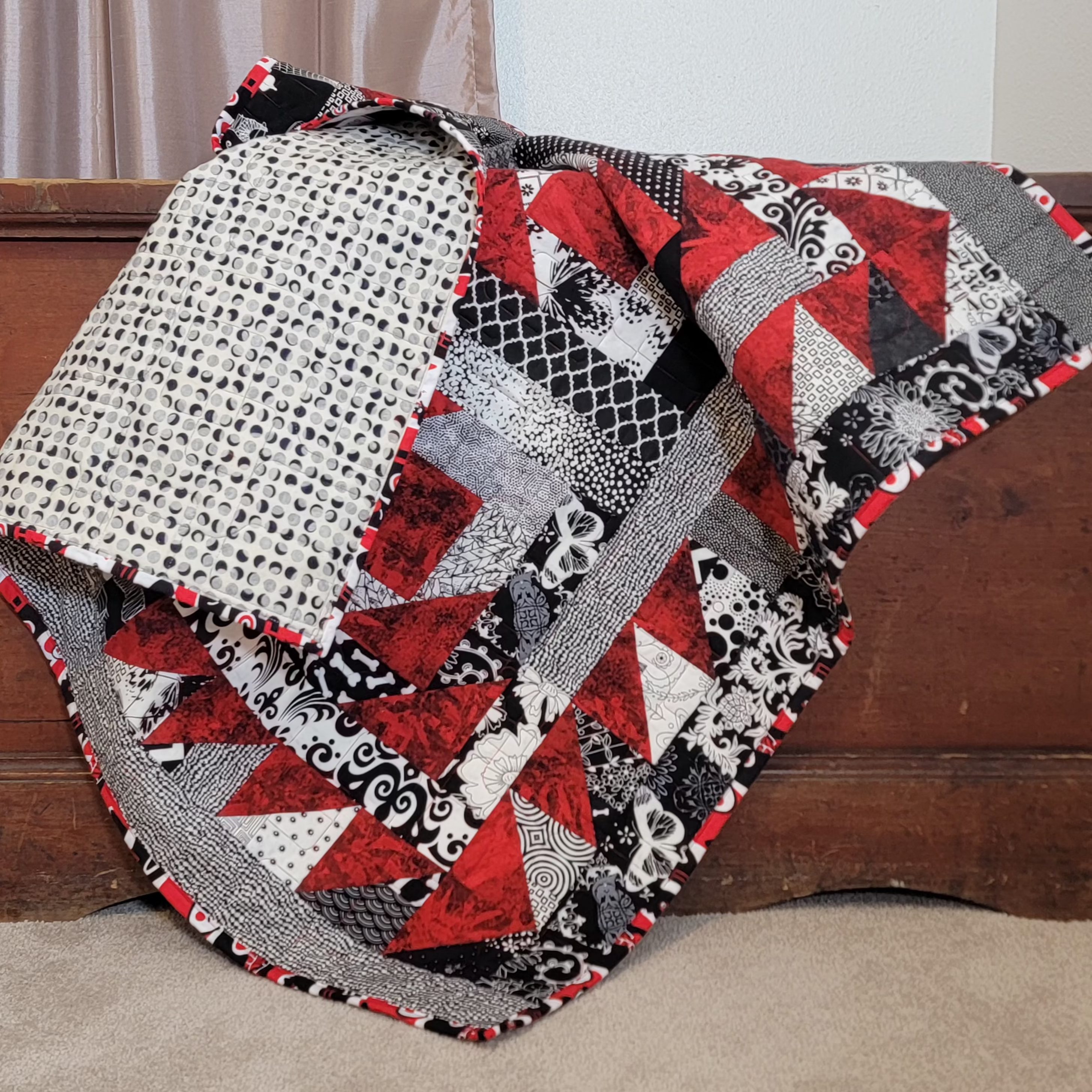 A red, white and black geometric quilt draped over a brown wooden chest, with a glimpse of the reverse side of the quilt in a black and white moon print. 