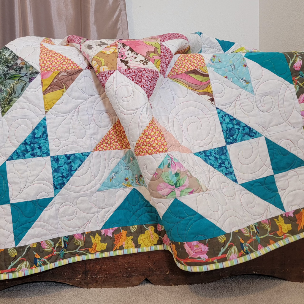 A handmade quilt in white, teal, orange and burgundy colours in multiple prints with a central star and a floral border draped over a wooden chest