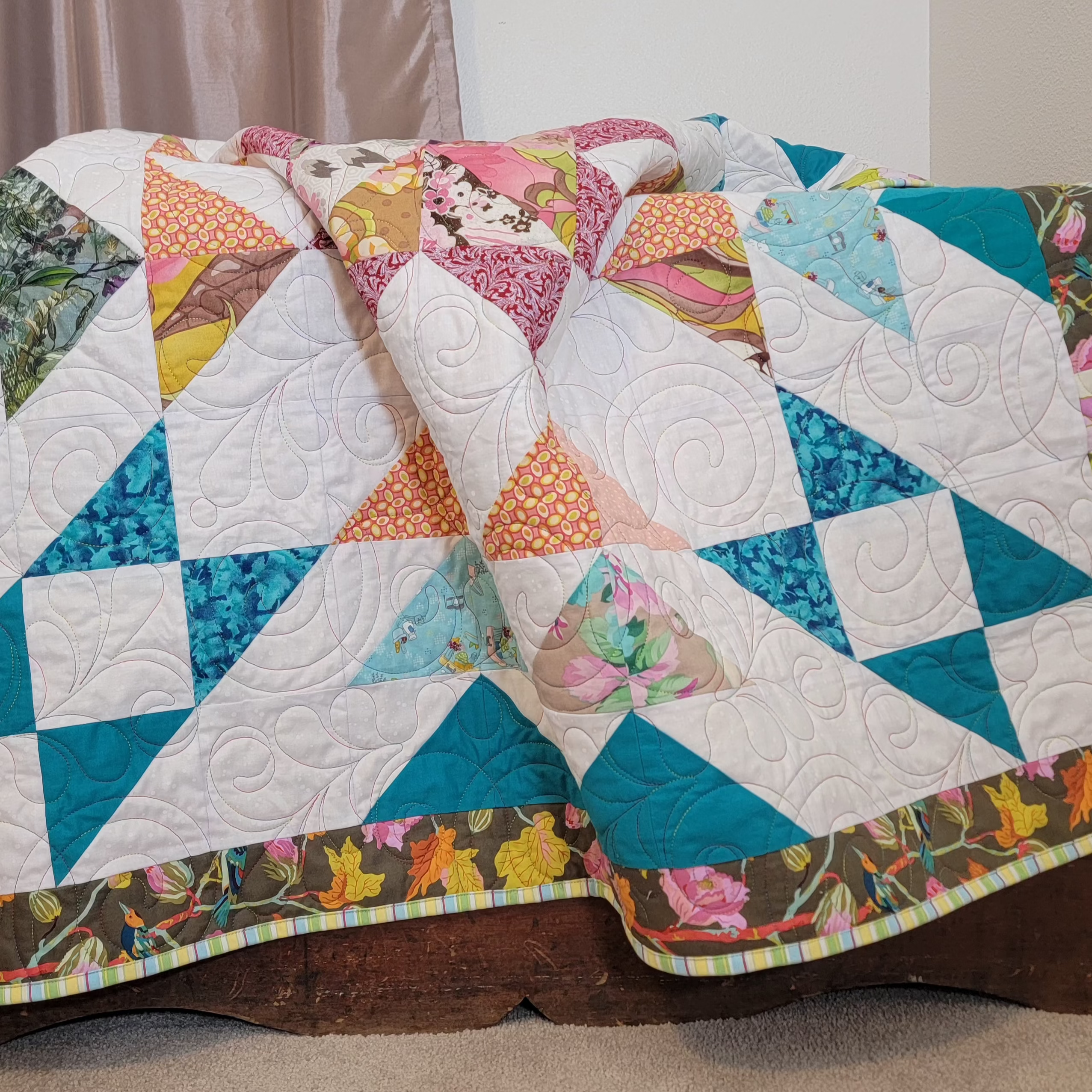 A handmade quilt in white, teal, orange and burgundy colours in multiple prints with a central star and a floral border draped over a wooden chest