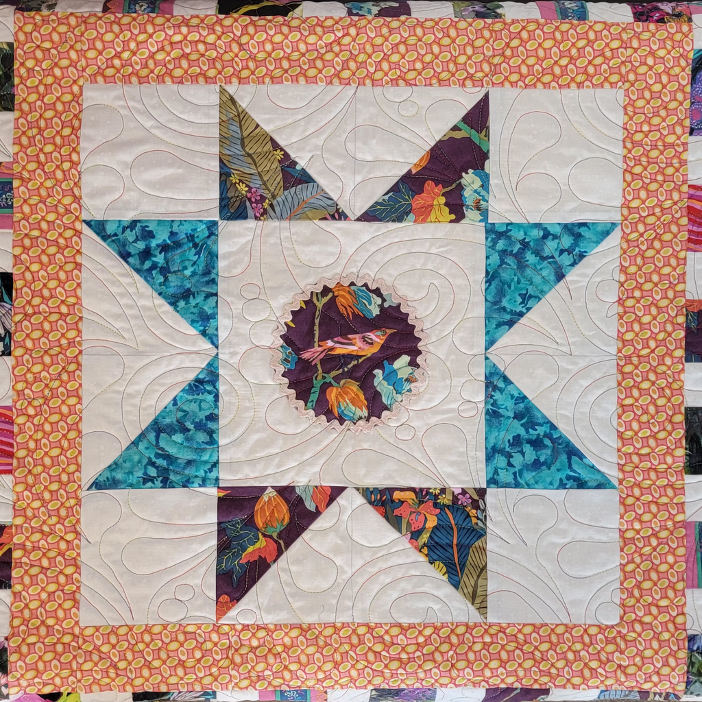 A square quilted textile featuring a central bird motif surrounded by teal and orange patterned star-like sections, with a peach floral border.