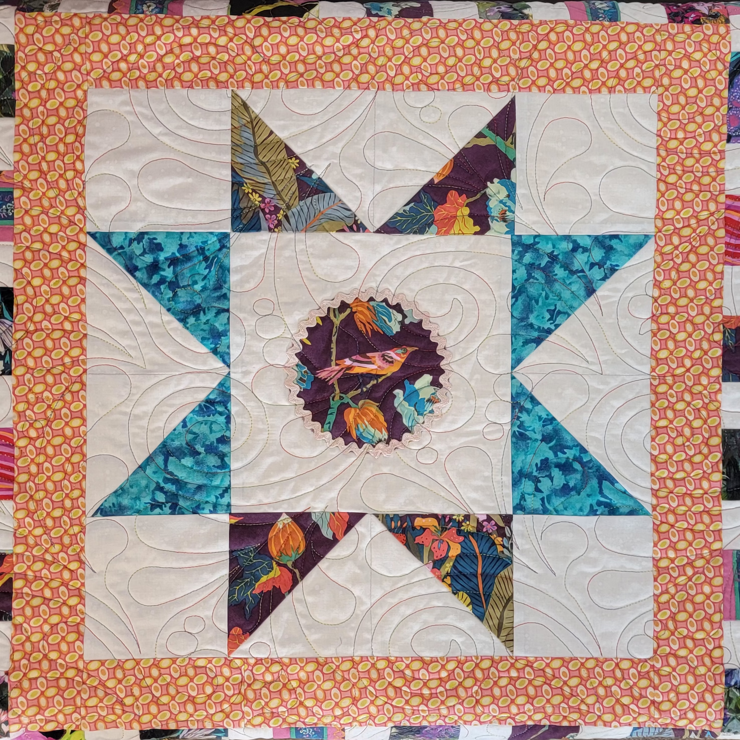 A square quilted textile featuring a central bird motif surrounded by teal and orange patterned star-like sections, with a peach floral border.