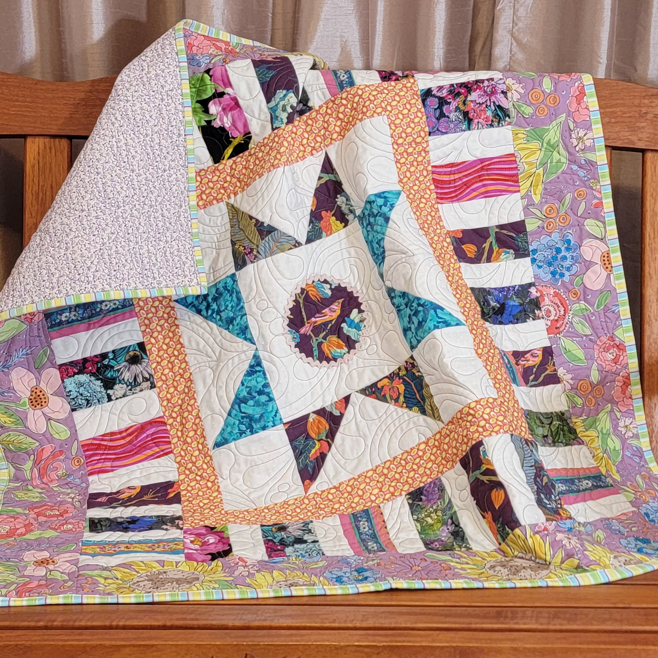 A colorful patchwork quilt draped over a wooden bench in front of beige curtains. The quilt features a central starburst pattern with blue, white, and orange sections, surrounded by floral and striped fabric borders.