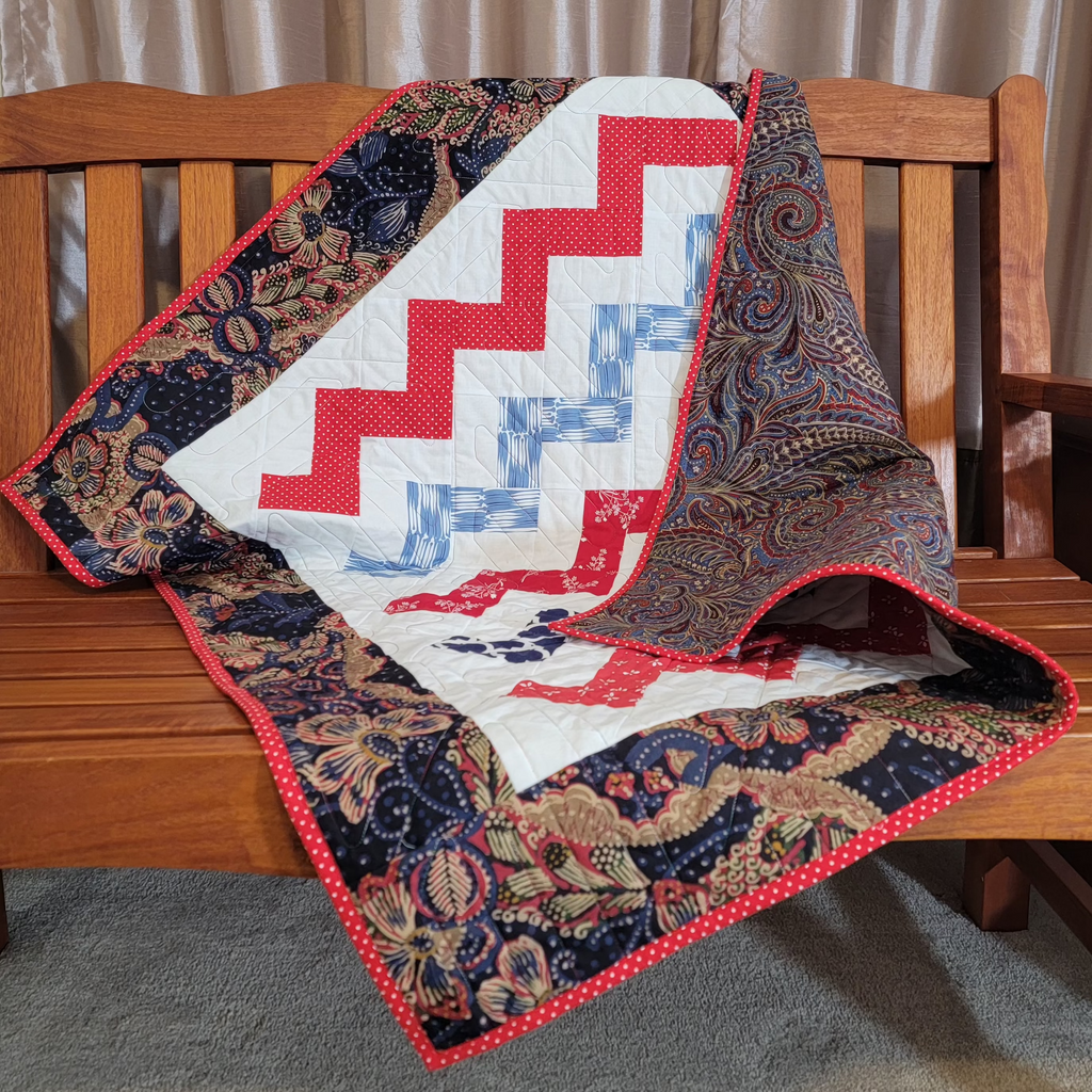A handmade quilted blanket with a white center featuring red and blue zigzag stripes, draped over a wooden bench with a dark floral paisley patterned border and red polka dot trim.