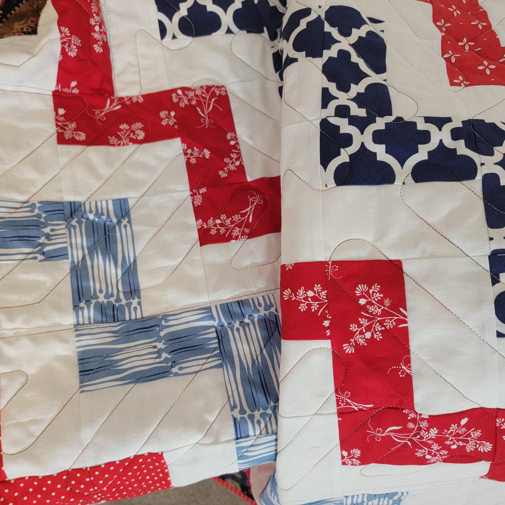 A close-up of a handmade quilt with a patriotic color scheme of red, white, and blue. The quilt features various patterned fabric sections including solid red, red with white floral prints, blue and white stripes, and navy blue with white Moroccan trellis patterns, all stitched together with white quilting lines.