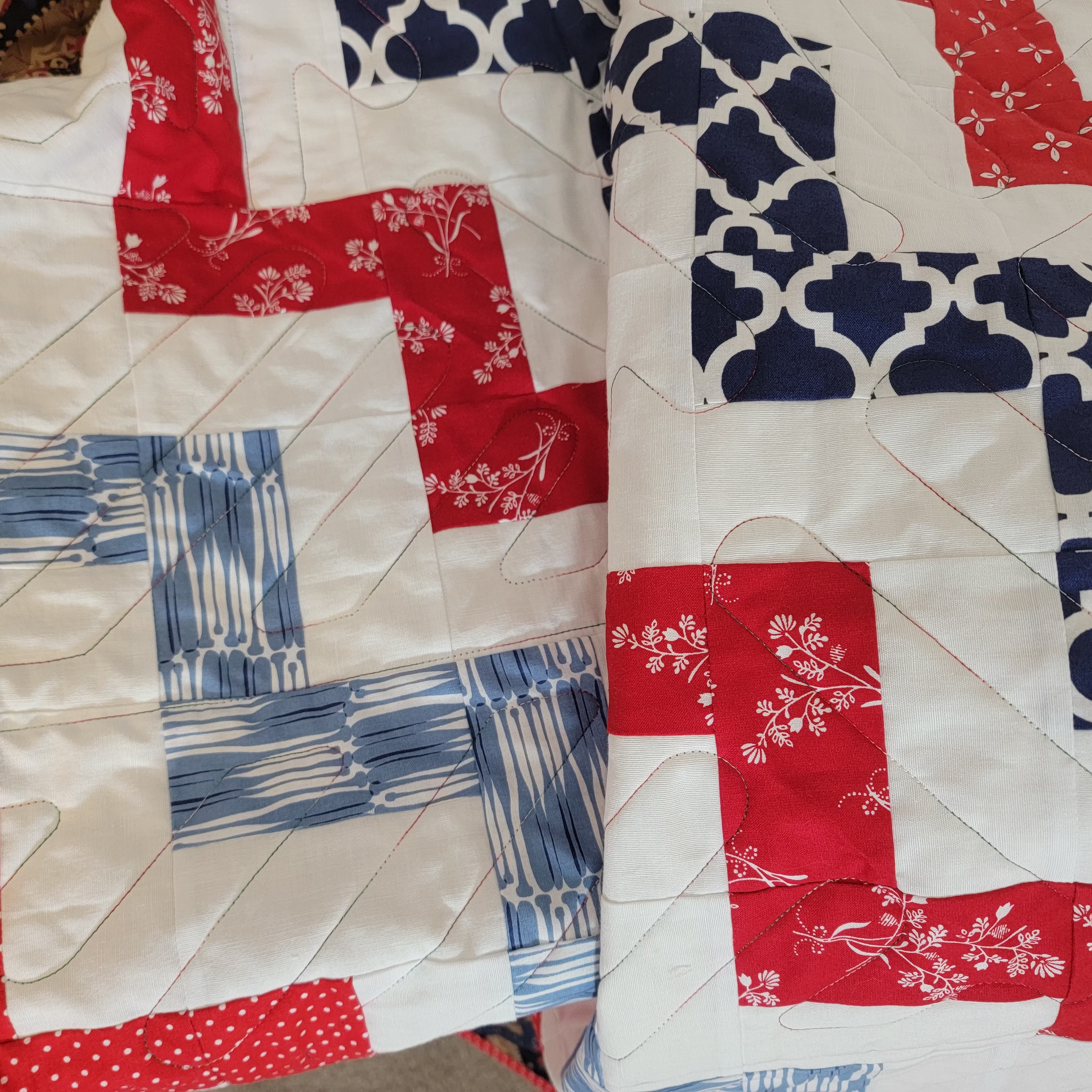 A close-up of a handmade quilt with a patriotic color scheme of red, white, and blue. The quilt features various patterned fabric sections including solid red, red with white floral prints, blue and white stripes, and navy blue with white Moroccan trellis patterns, all stitched together with white quilting lines.