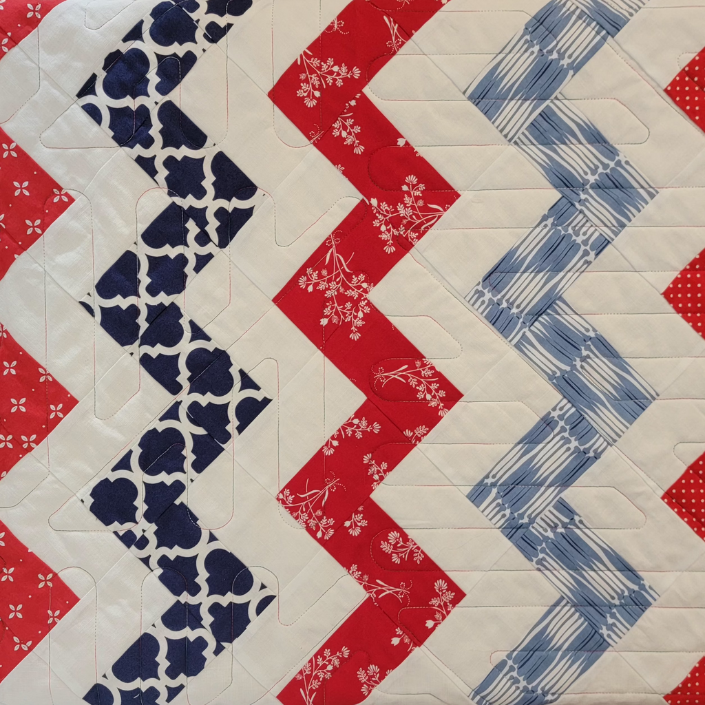 A close-up of a quilted fabric featuring a chevron pattern with alternating zigzag stripes in red, navy blue, and light blue on a white background.