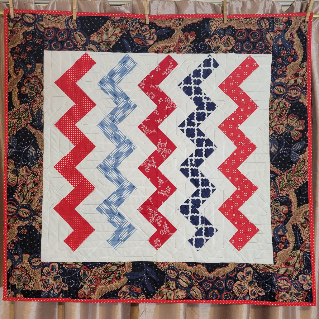 A handmade quilt hanging from a wooden dowel with clothespins against a beige curtain backdrop. The quilt features a central white square with four vertical zigzag stripes in red, light blue, and dark navy patterns, surrounded by a dark floral patterned border with red piping.