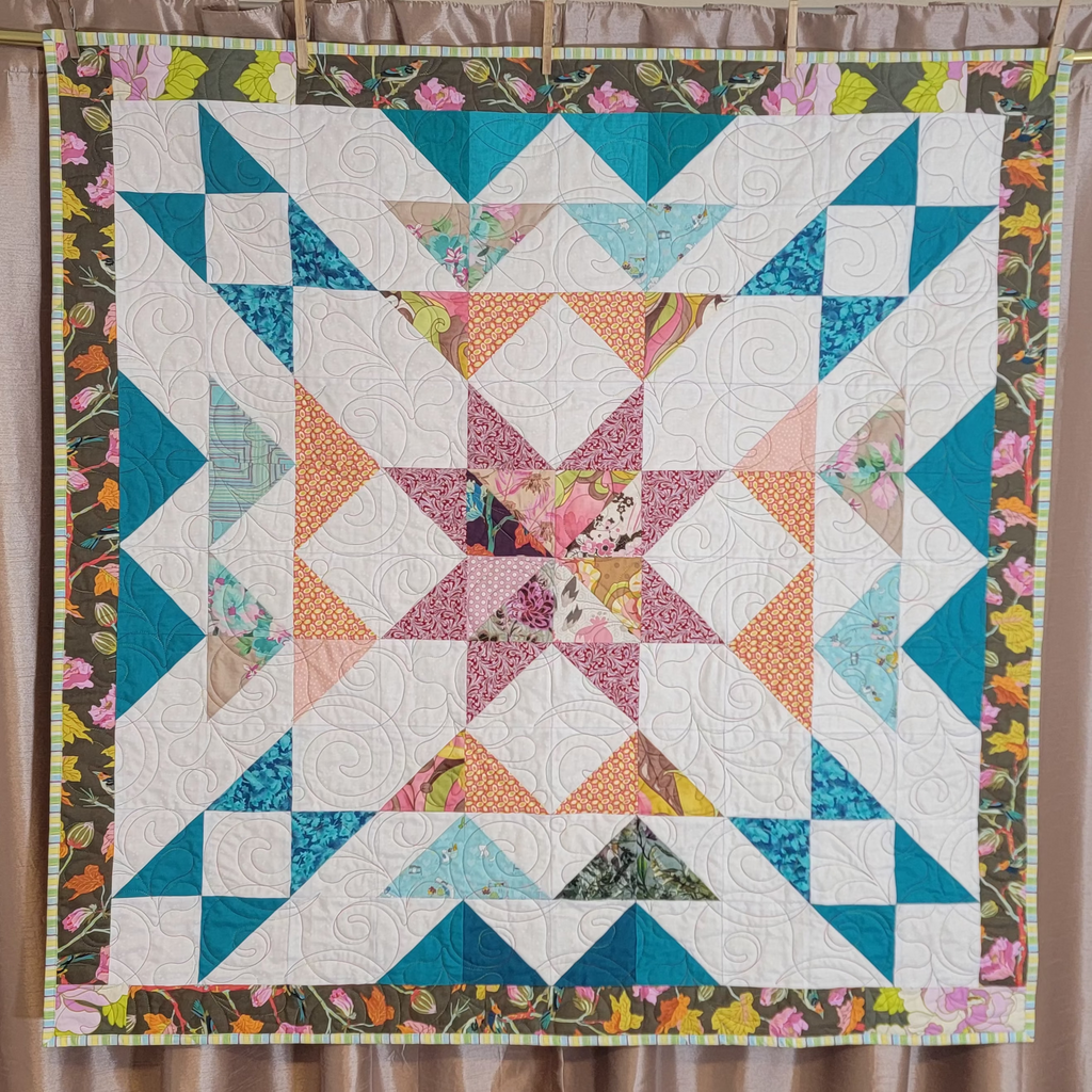 A handmade quilt in white, teal, orange and burgundy colours in multiple prints with a central star and a floral border hung up on a gold rail with a dusky pink background. 