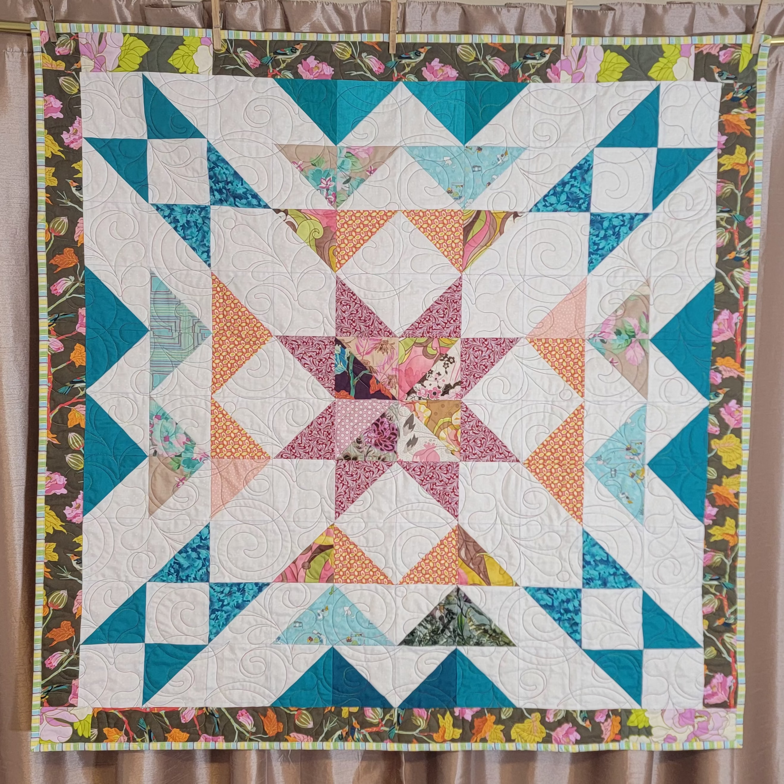 A handmade quilt in white, teal, orange and burgundy colours in multiple prints with a central star and a floral border hung up on a gold rail with a dusky pink background. 