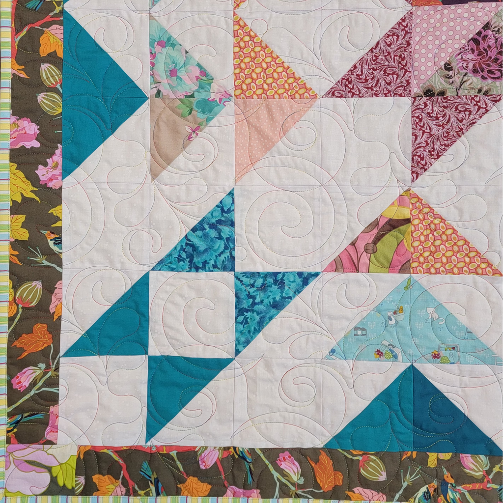 A close up of a handmade quilt with various triangle section in various prints in blue, orange and burgundy. All with a floral border. 