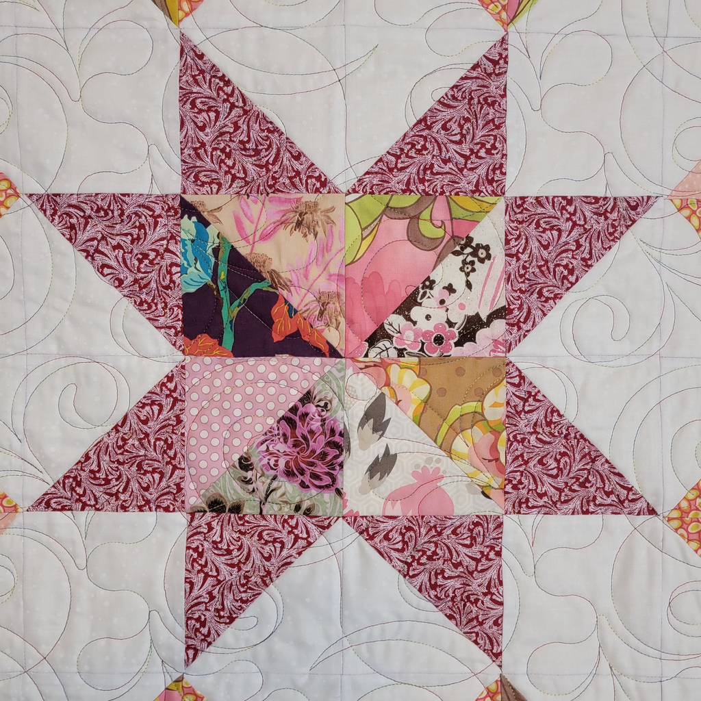A close up of a handmade quilt with a central star design. The star is made up of a burgundy leaf print with 4 square central blocks in multiple prints. The star is surrounded by a white background with intricate stitching details. 