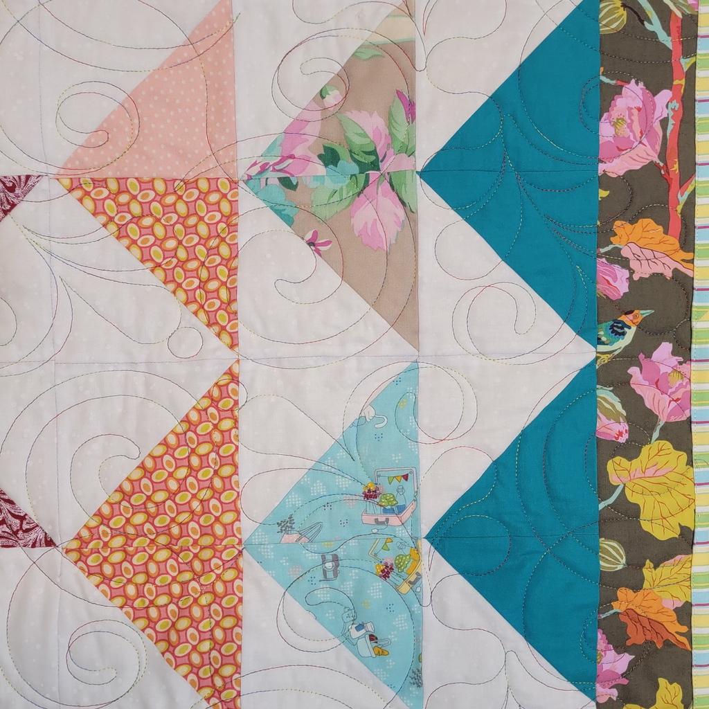 A close up of a handmade quilt featuring triangle sections in blue and orange prints with a floral border with a white background with intricate stitching details.