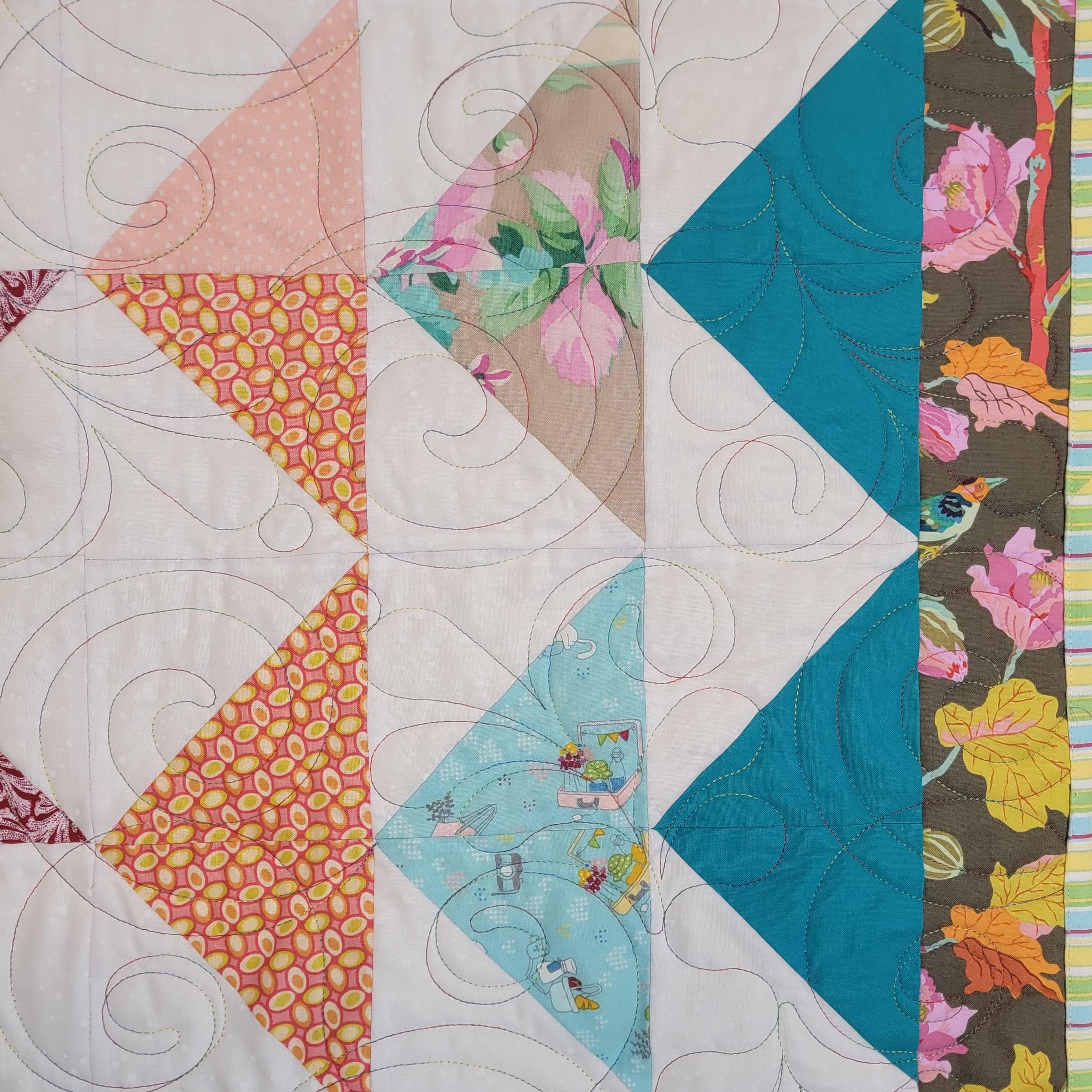 A close up of a handmade quilt featuring triangle sections in blue and orange prints with a floral border with a white background with intricate stitching details.