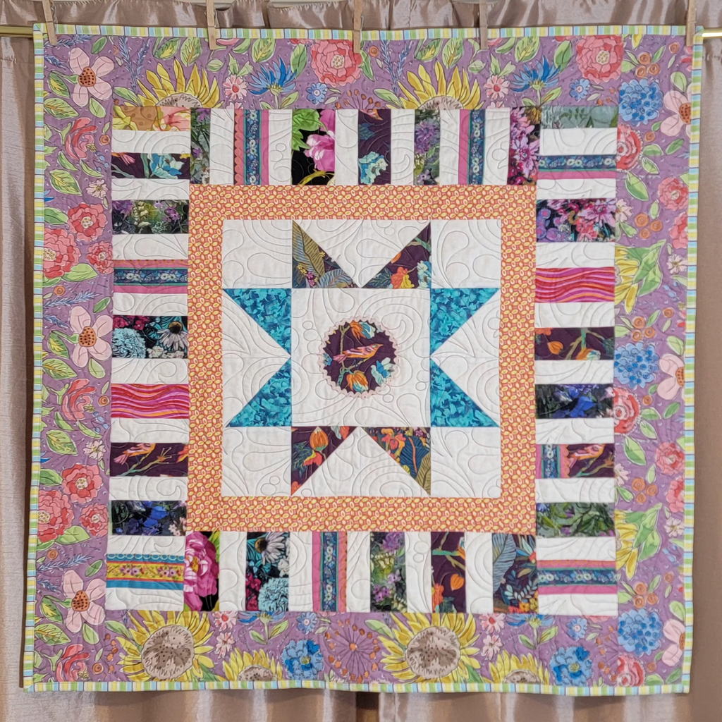 A colourful patchwork quilt with a star in the centre surrounded by striped floral prints and bordered with a large floral print in purple hung up on a gold rail.
