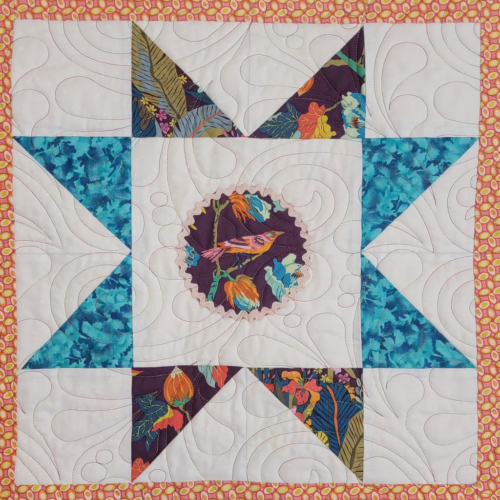 A close-up of a colorful quilted fabric featuring a geometric starburst pattern with white quilted sections, teal patterned triangles, and a central circular motif with a bird.