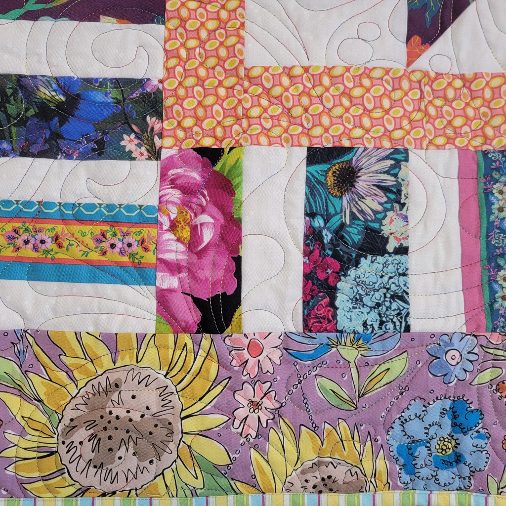 A close-up of a colorful quilted fabric panel with floral and botanical patterns, including pink flowers, sunflowers, butterflies, and striped borders on purple, pink, yellow, blue, and white fabrics.