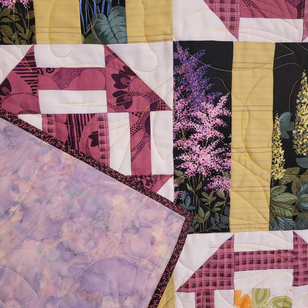 Close up of a quilt with multiple floral and garden inspired prints in different shapes. The reverse of the quilt is visible and is a purple and blue hand dyes batik.