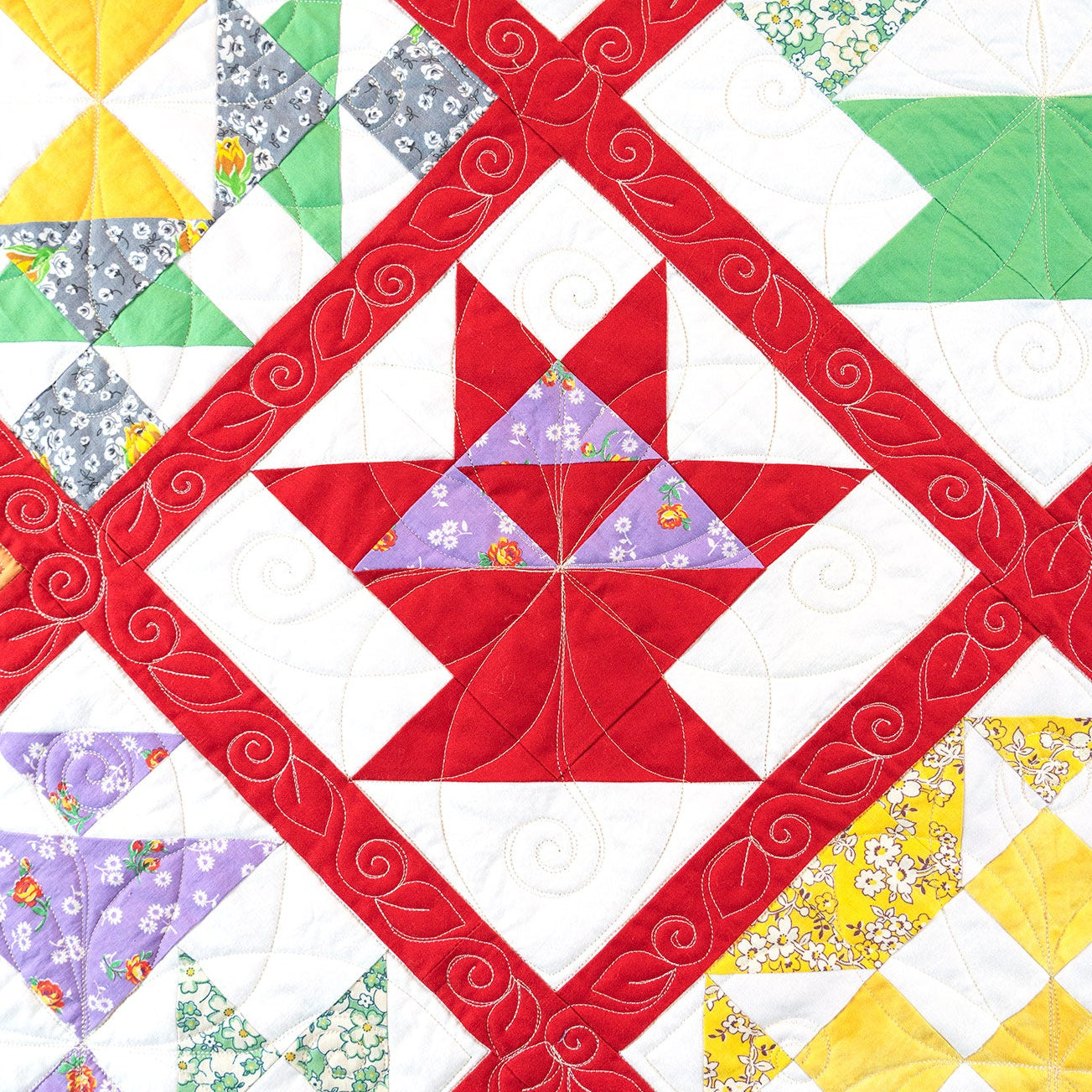 A close-up of a quilted fabric featuring a geometric patchwork design with red diagonal bands, a central red and purple star motif, and surrounding blocks in green, yellow, orange, and patterned prints on white.