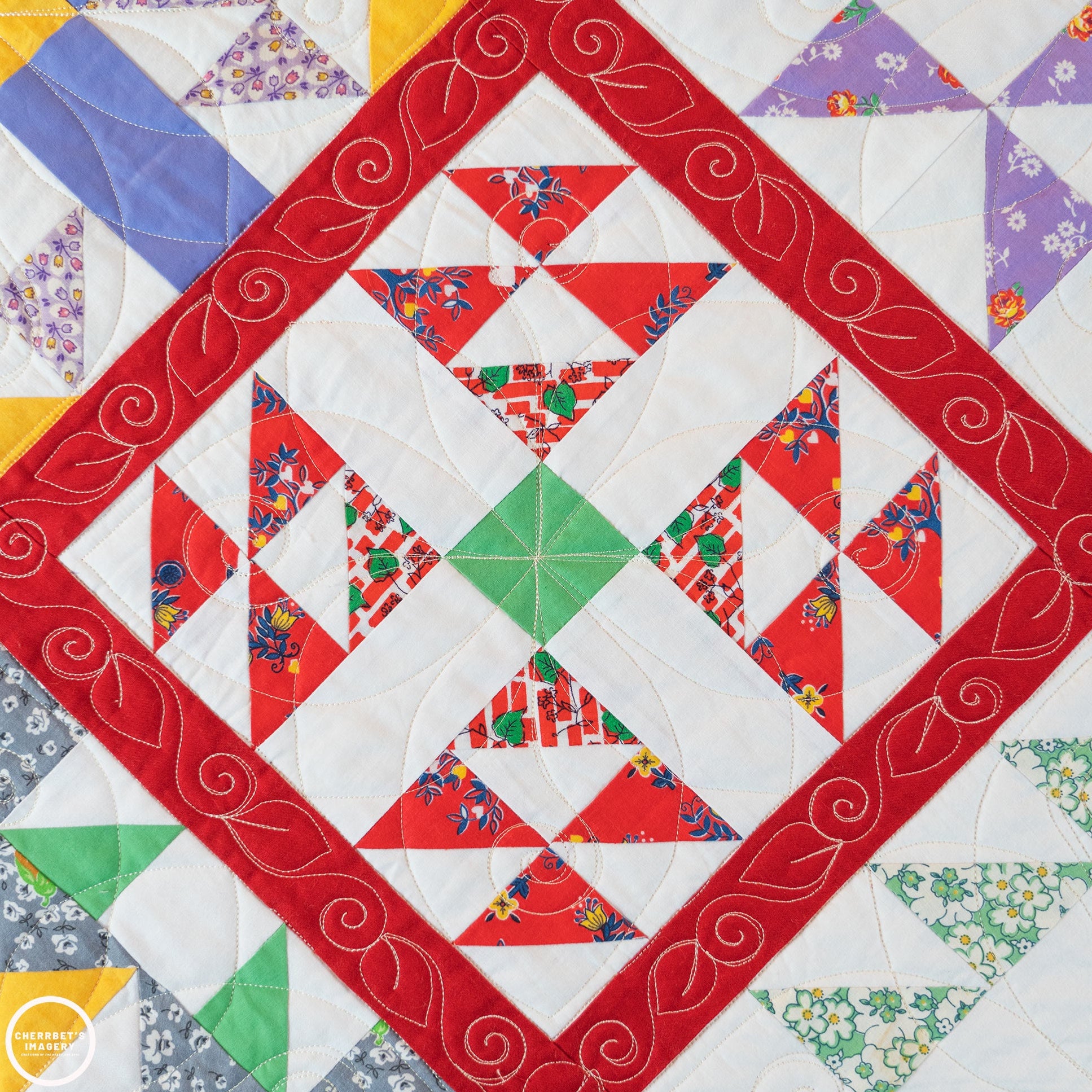 A close-up of a quilted textile with a geometric patchwork design. The pattern features a wide red border with white swirling embroidery, red and white triangular sections, a green center diamond, and smaller floral fabric patches in blue, purple, green, and gray around the edges.