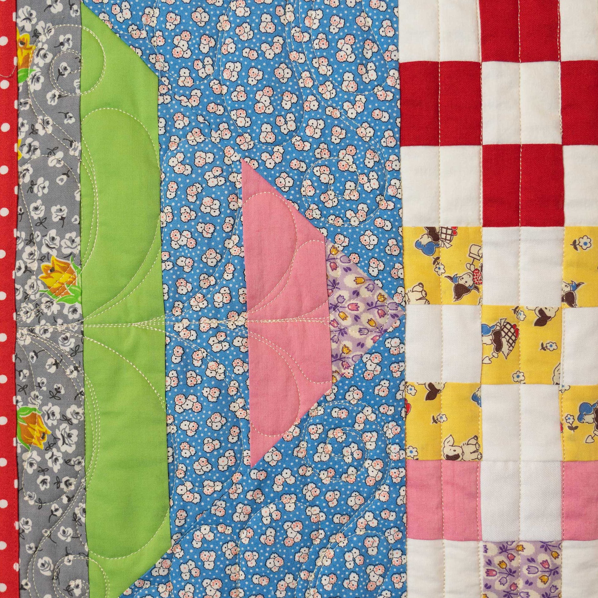 A close-up of a handmade quilted fabric panel featuring multiple colorful patchwork sections, including a blue floral center, a green curved panel, a pink triangular panel, and a red-and-white checkered section on the right.