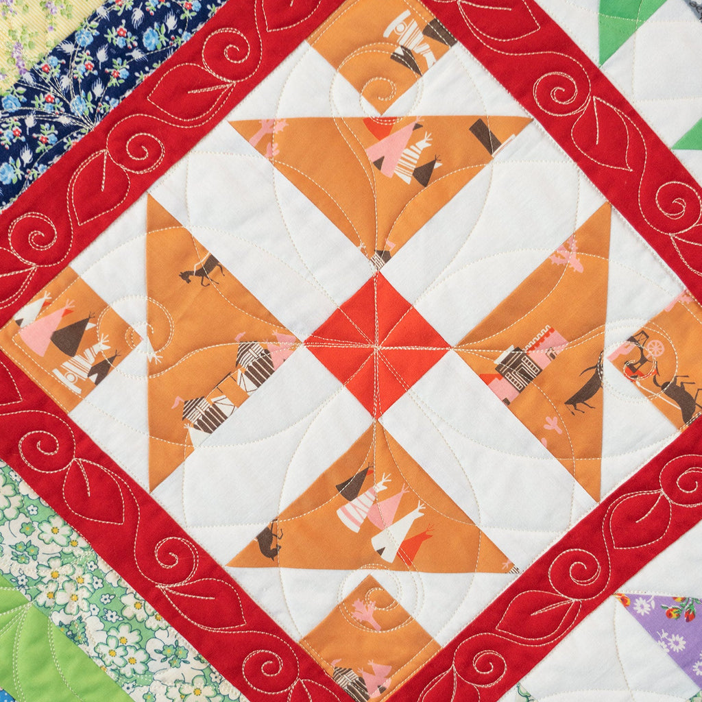 A close-up of a quilted fabric design featuring a central orange and white geometric star pattern surrounded by a red border with white swirl embroidery, with other colorful floral and striped fabric borders visible around the edges.