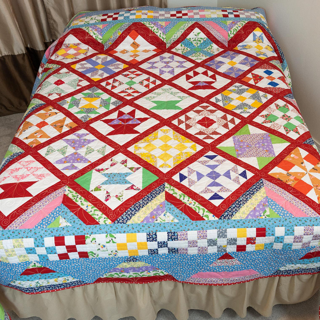 A colorful patchwork quilt draped over a bed with a beige ruffled bed skirt, photographed in a bedroom setting with curtains and carpet visible.