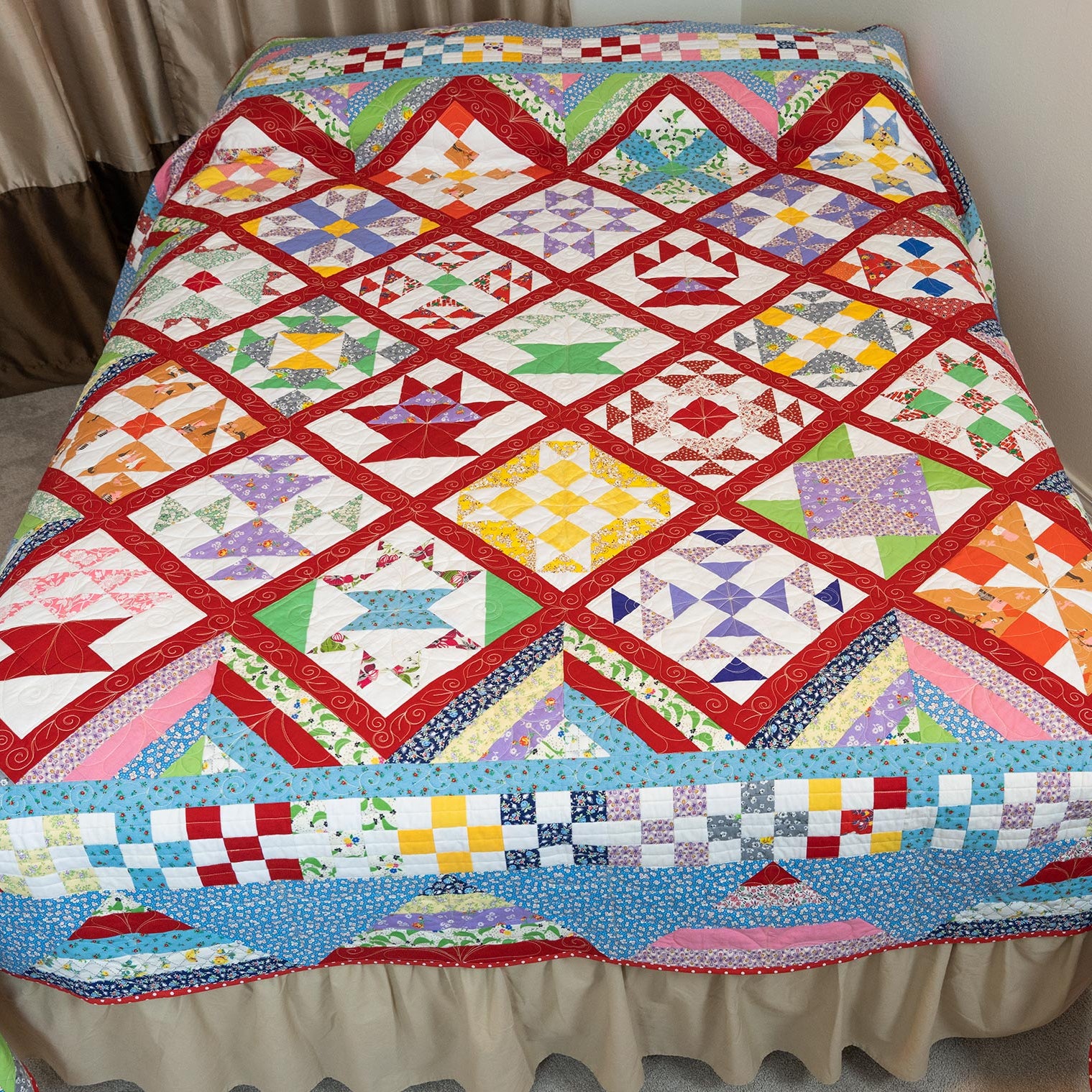 A colorful patchwork quilt draped over a bed with a beige ruffled bed skirt, photographed in a bedroom setting with curtains and carpet visible.