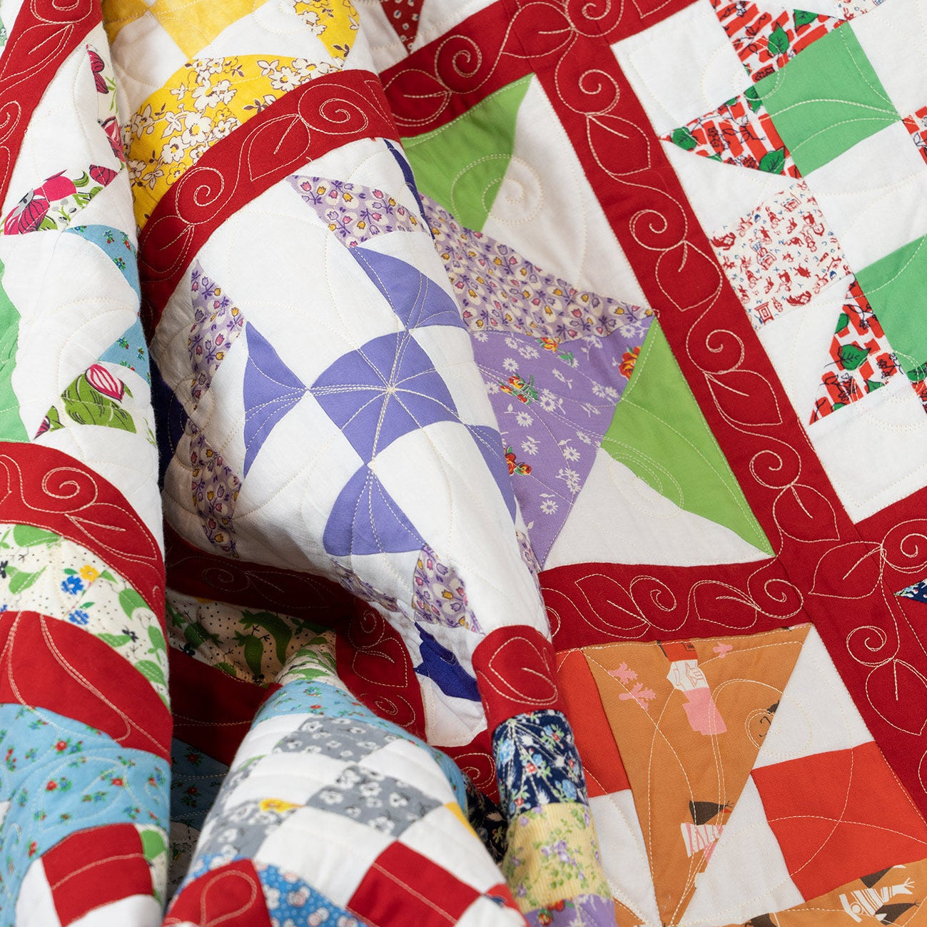 A close-up of a colorful quilted fabric with patchwork blocks, red borders, and swirling stitched details.