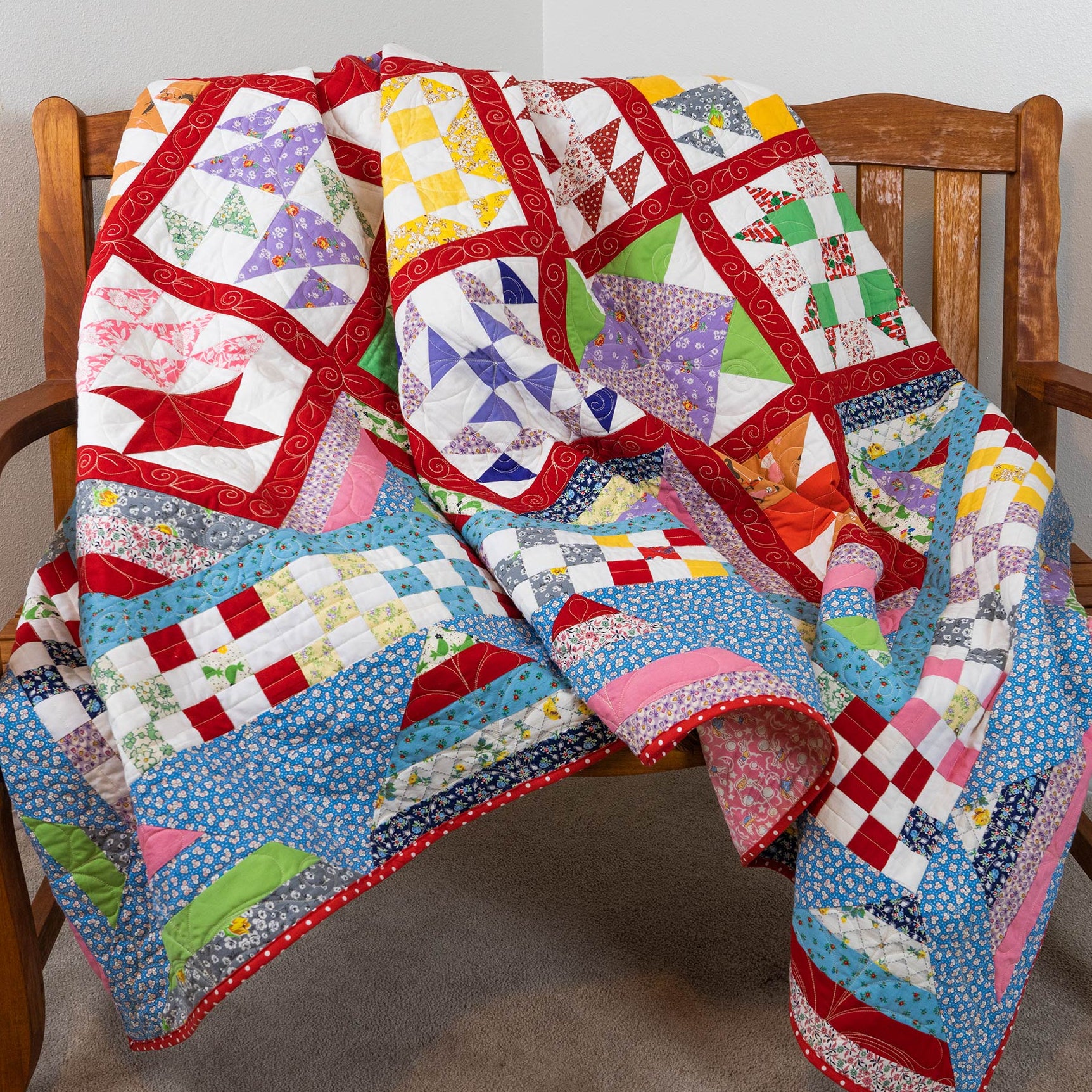 A colorful patchwork quilt draped over a wooden armchair in a home setting, featuring various geometric and floral patterns in red, blue, pink, yellow, green, and white.