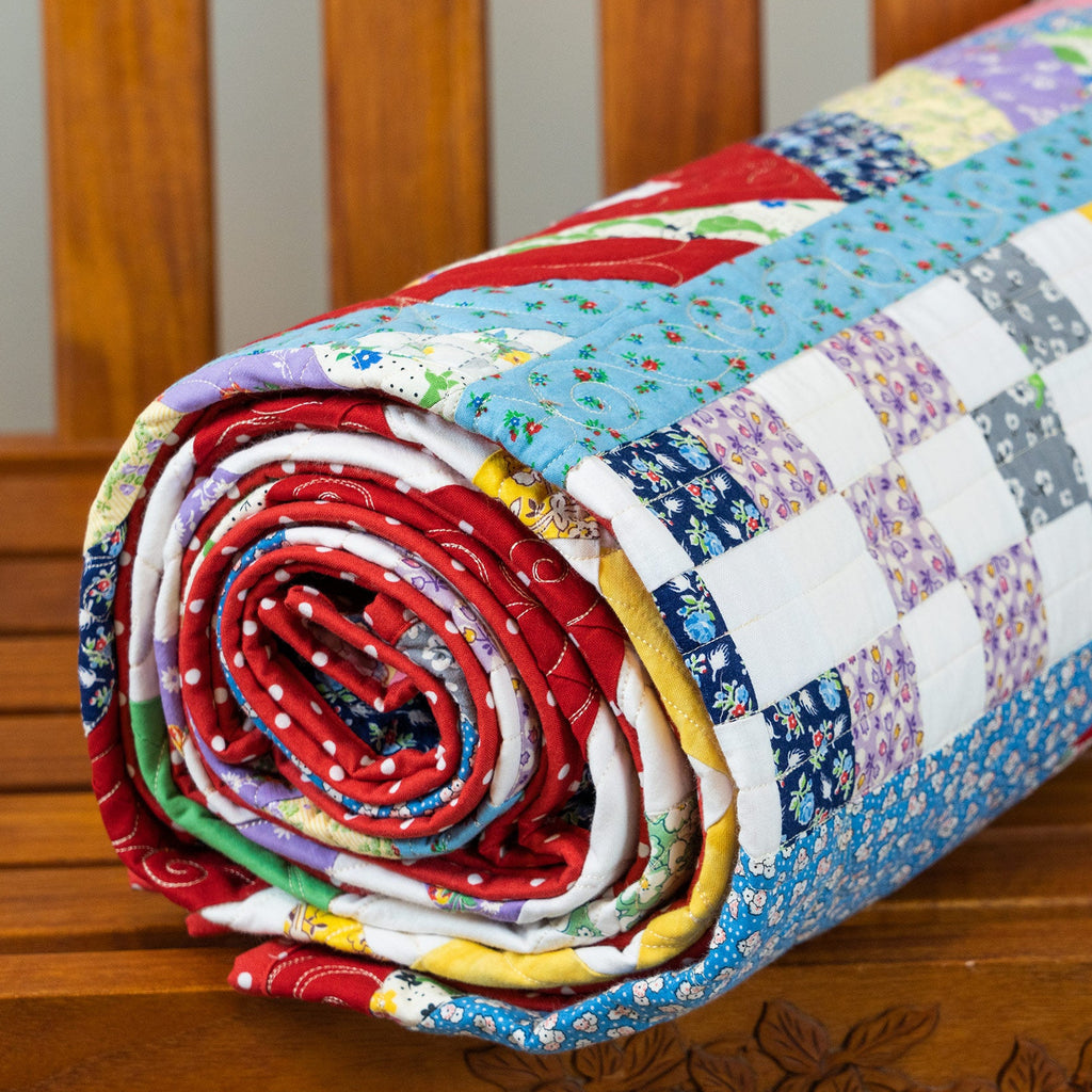 A rolled quilt with a colorful patchwork design featuring floral, checkered, and solid fabric blocks, displayed on a carved wooden bench.