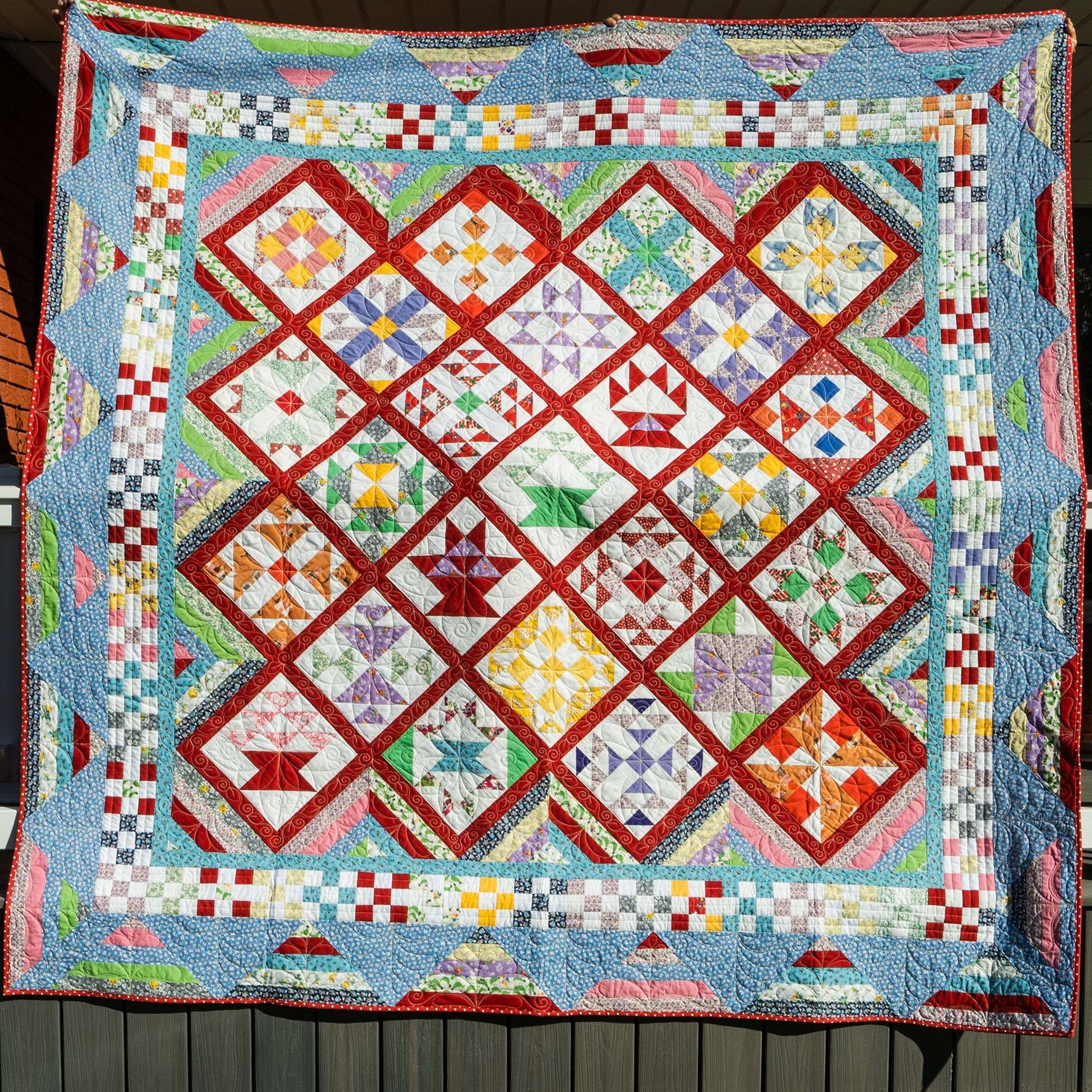 A large handmade quilted patchwork blanket or quilt hanging outdoors on a porch. The design features a complex diamond lattice pattern with multiple colors including red, blue, green, yellow, orange, and white, bordered by a blue and white checkered pattern.