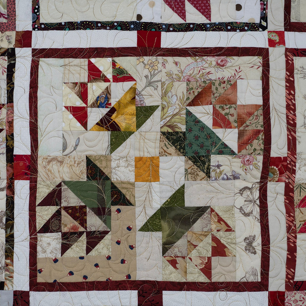 A close-up of a handmade quilt featuring a patchwork design with various geometric and floral blocks in muted red, green, beige, and brown tones, framed by a dark red border.