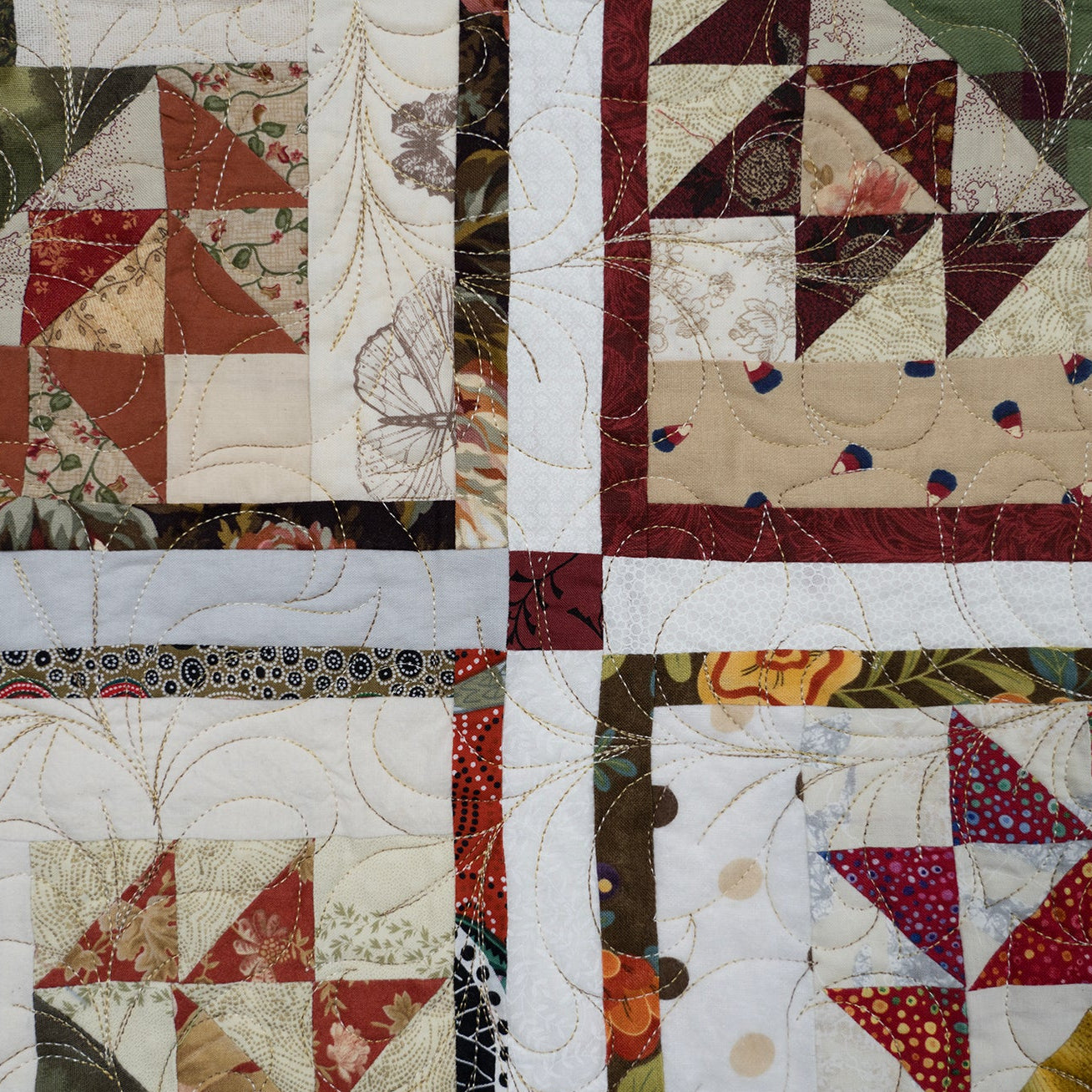 A close-up of a quilted patchwork textile with four quadrants, featuring floral and geometric fabric blocks in muted reds, creams, greens, and browns, with visible quilting stitches and a white cross-shaped seam dividing the center.