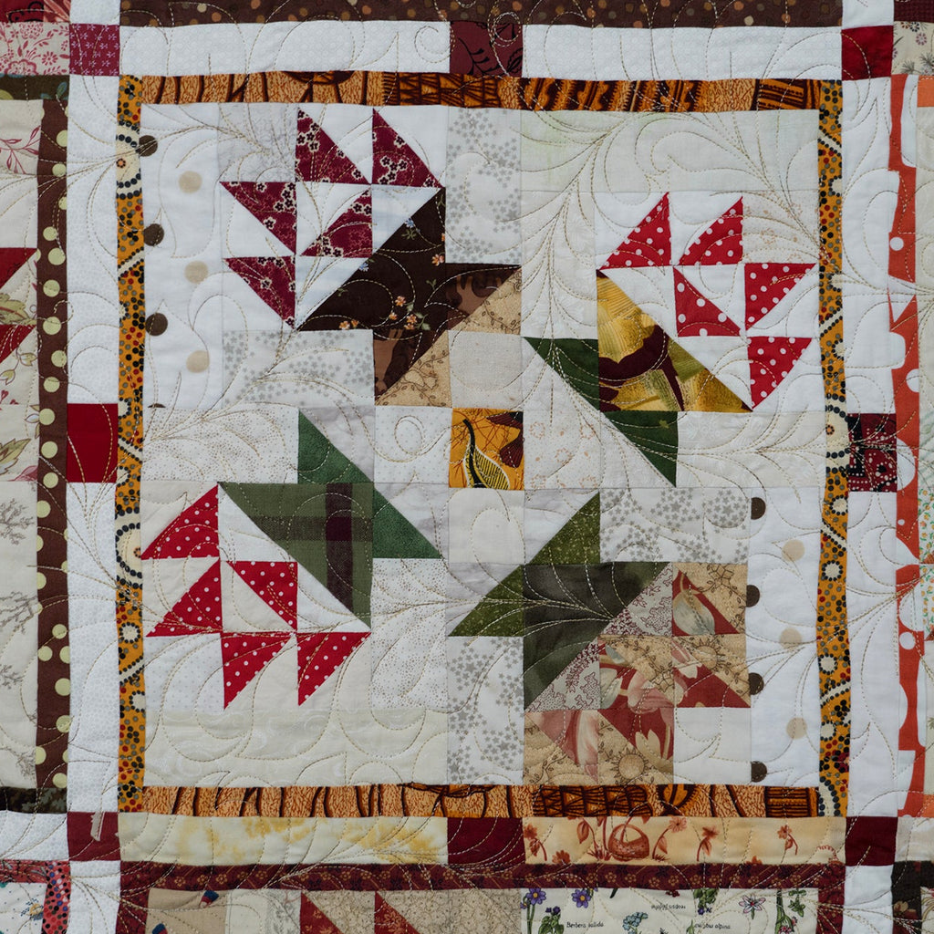 A close-up of a quilted textile featuring multiple patchwork panels with geometric and starburst patterns in red, beige, brown, green, and white fabrics.