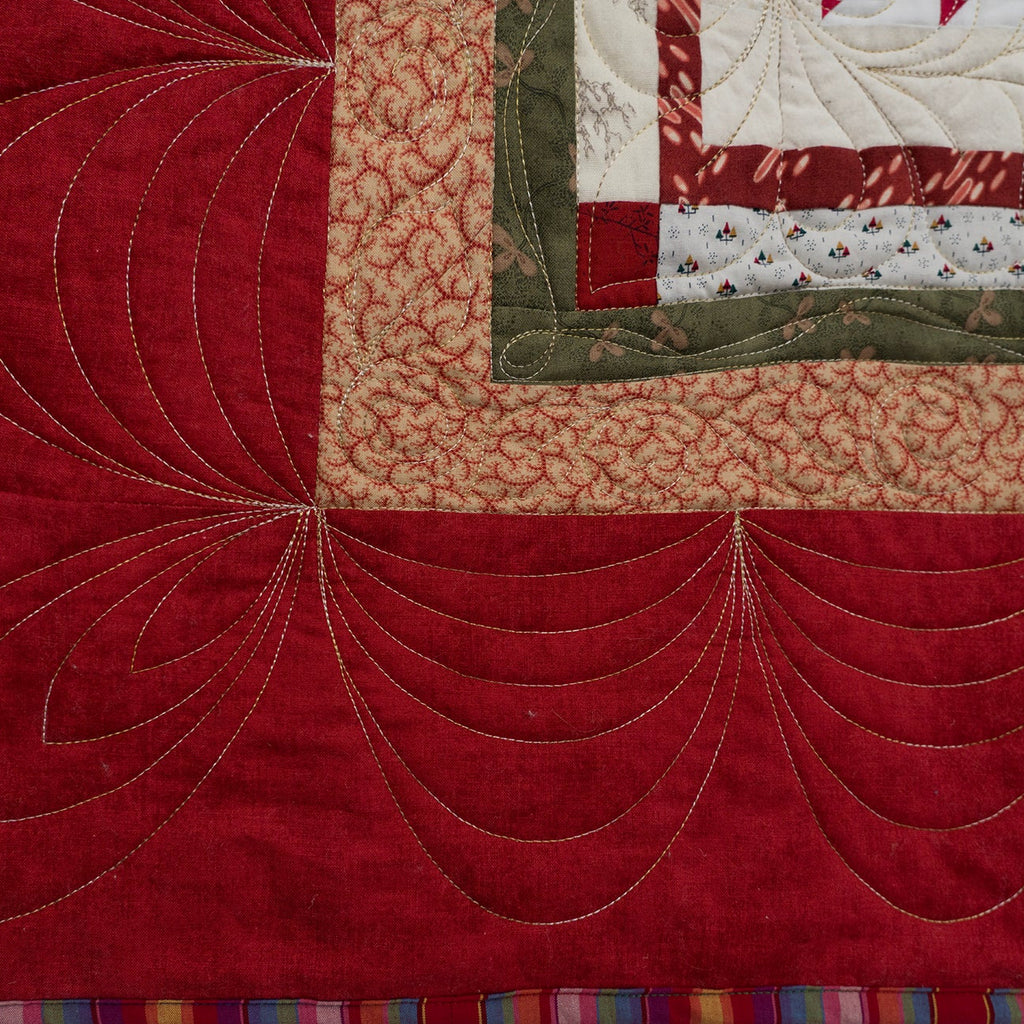 Close-up of a quilted textile showing a large red field with decorative curved stitching, a beige patterned section, a green border, and a multicolored striped edge.