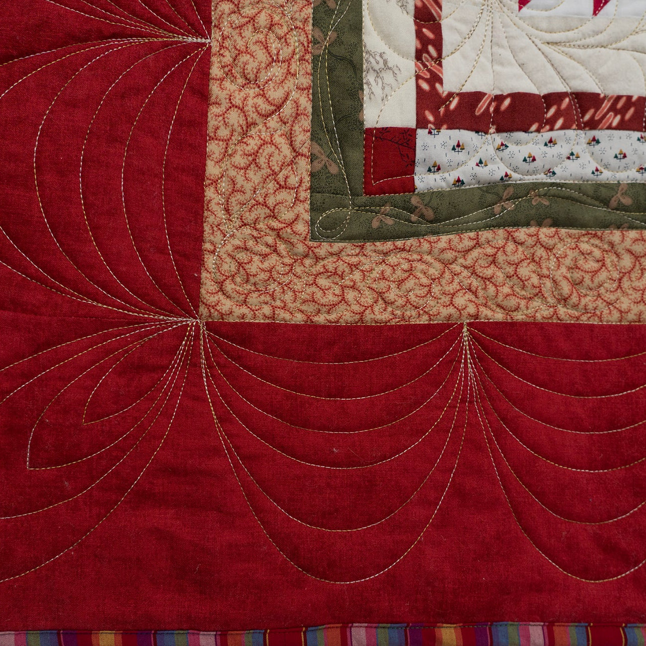 Close-up of a quilted textile showing a large red field with decorative curved stitching, a beige patterned section, a green border, and a multicolored striped edge.