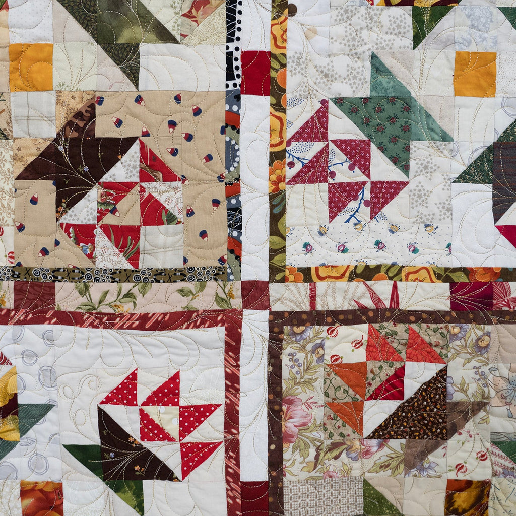 A square quilted textile featuring a four-quadrant patchwork design with geometric and floral blocks in red, green, beige, brown, and white fabrics.