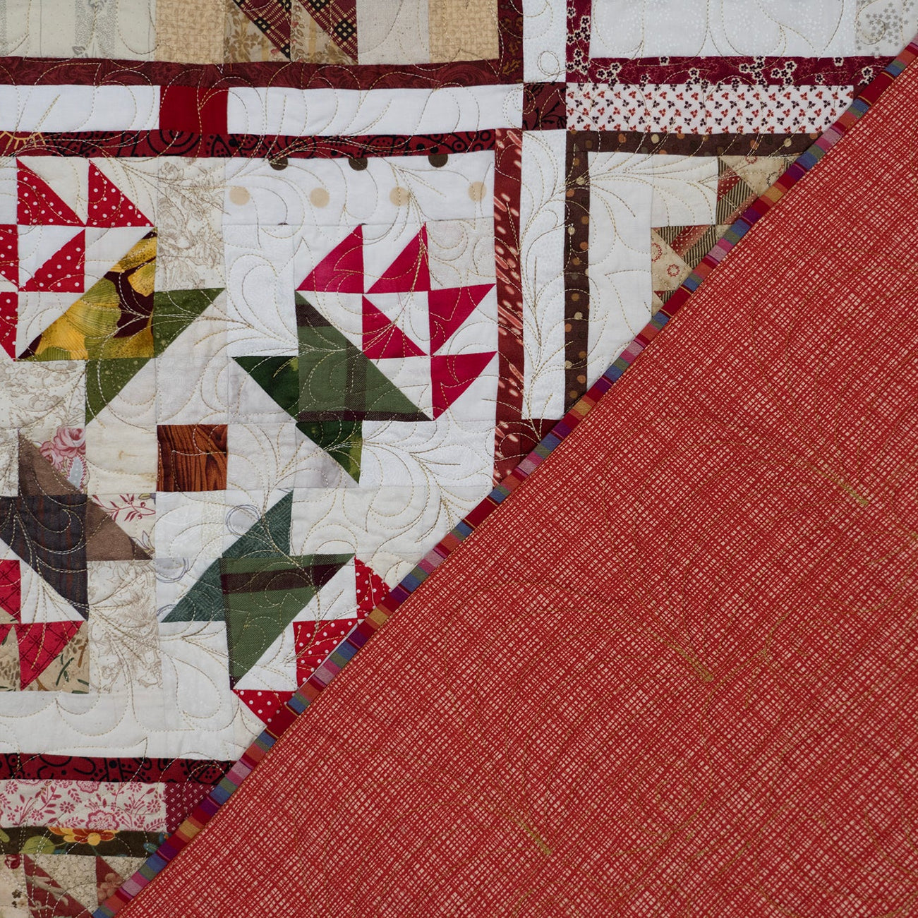 A close-up of a quilted textile with a diagonal split showing a red woven fabric section on one side and a patchwork section on the other. The patchwork includes cream, red, green, brown, and yellow fabric blocks.