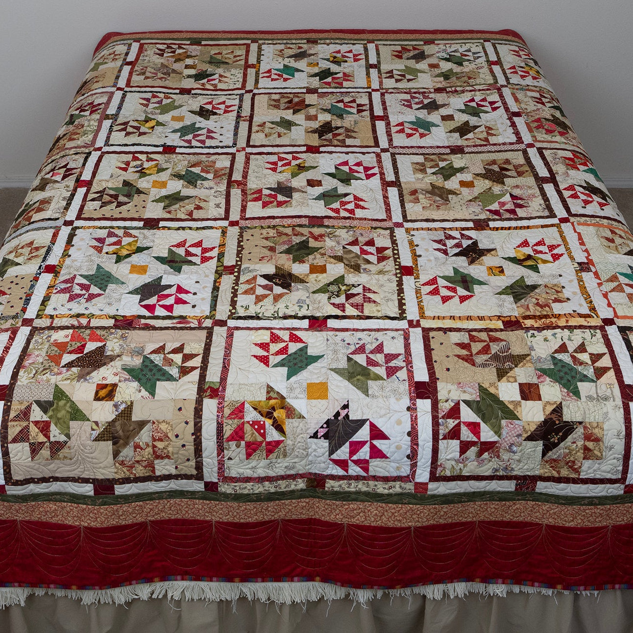 A vintage-style patchwork quilt with a geometric block pattern, draped over a bed with a beige ruffled bed skirt in a simple bedroom setting.
