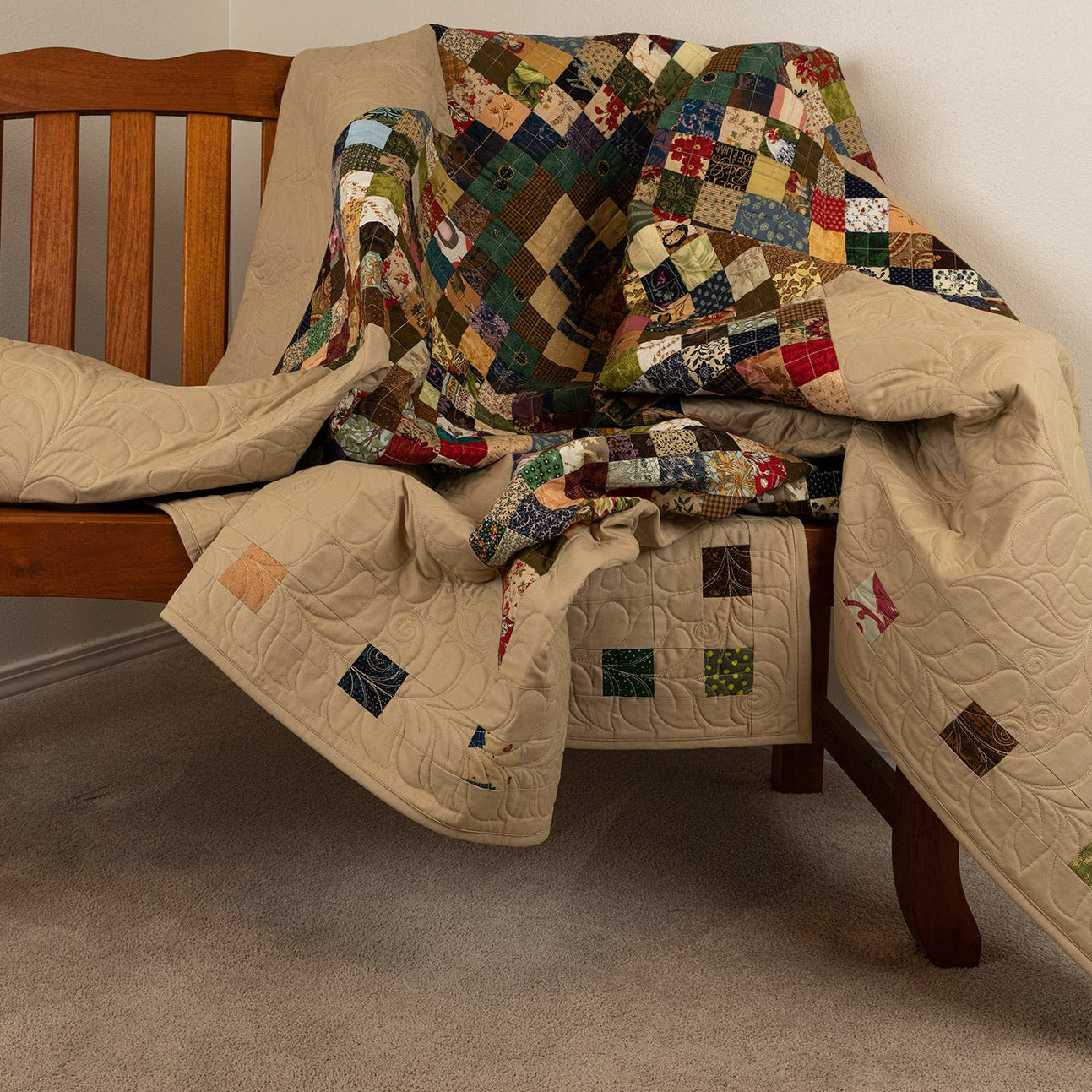 Wooden bench with a patchwork quilt draped over it against a plain wall.