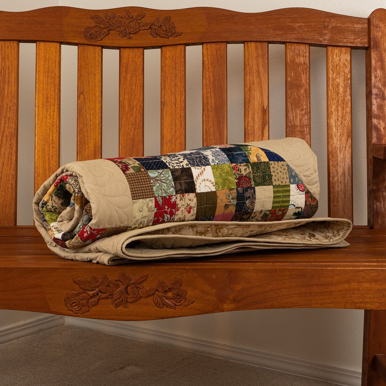 Wooden bench with a patchwork quilt draped over it against a plain wall.