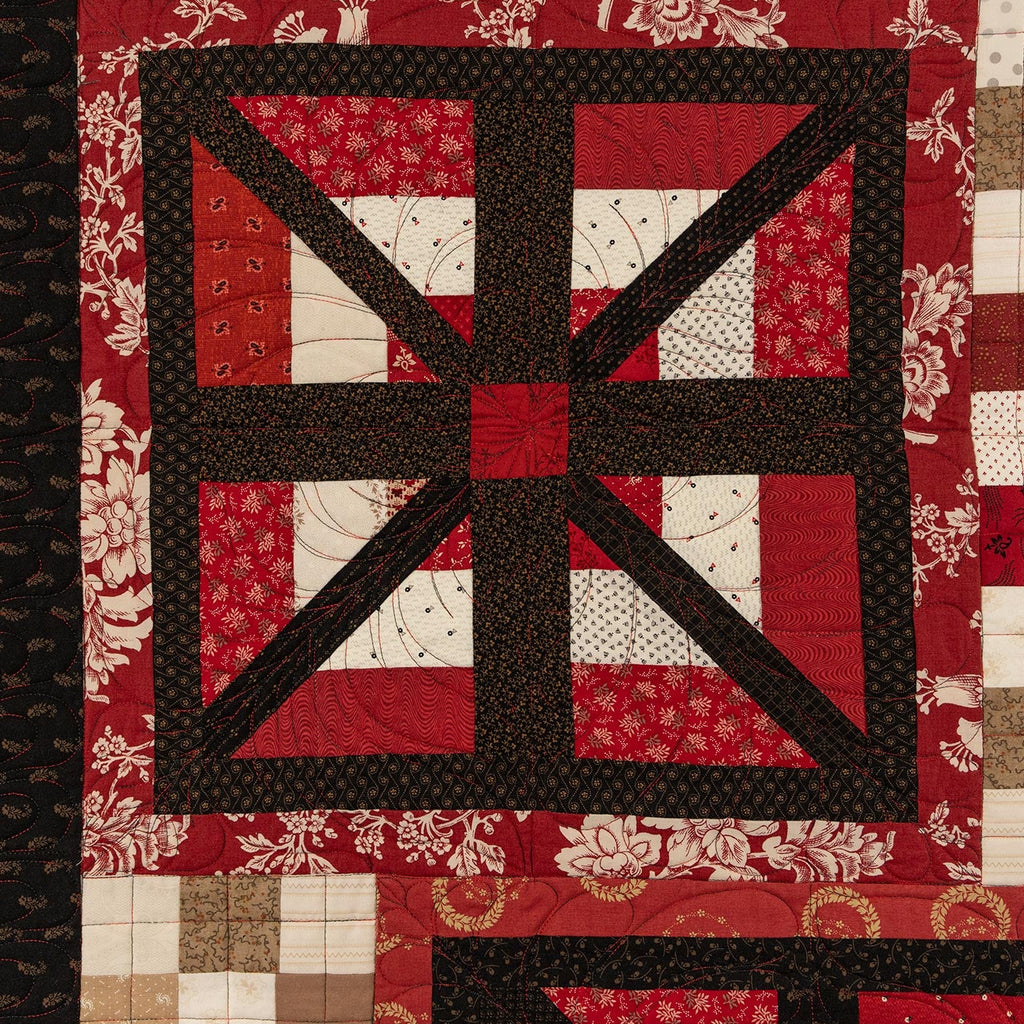 A close-up of a quilted textile with a geometric patchwork design in red, black, white, and beige tones. The central motif features a large black cross over red and white patterned sections, bordered by patchwork squares in red, beige, and white.