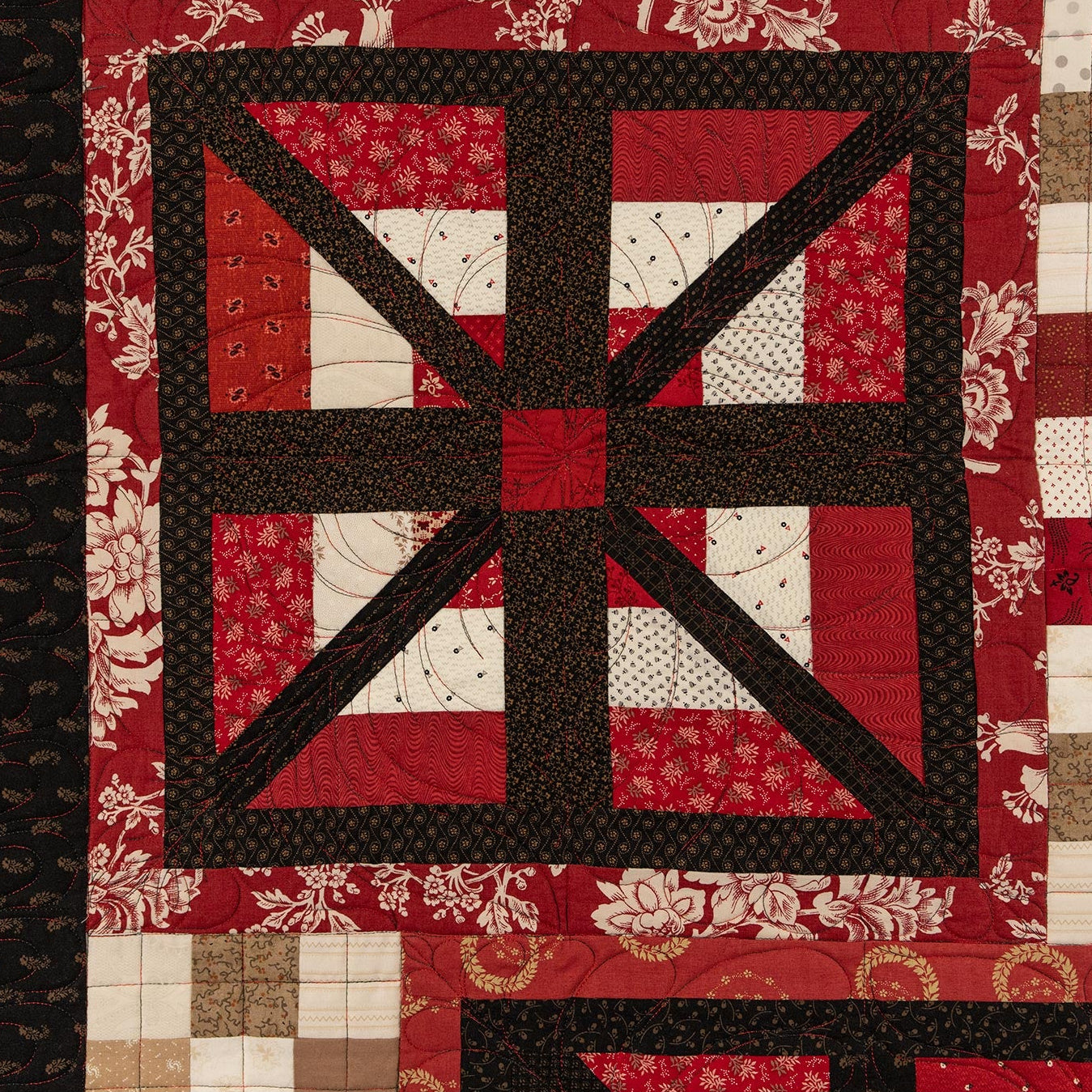 A close-up of a quilted textile with a geometric patchwork design in red, black, white, and beige tones. The central motif features a large black cross over red and white patterned sections, bordered by patchwork squares in red, beige, and white.