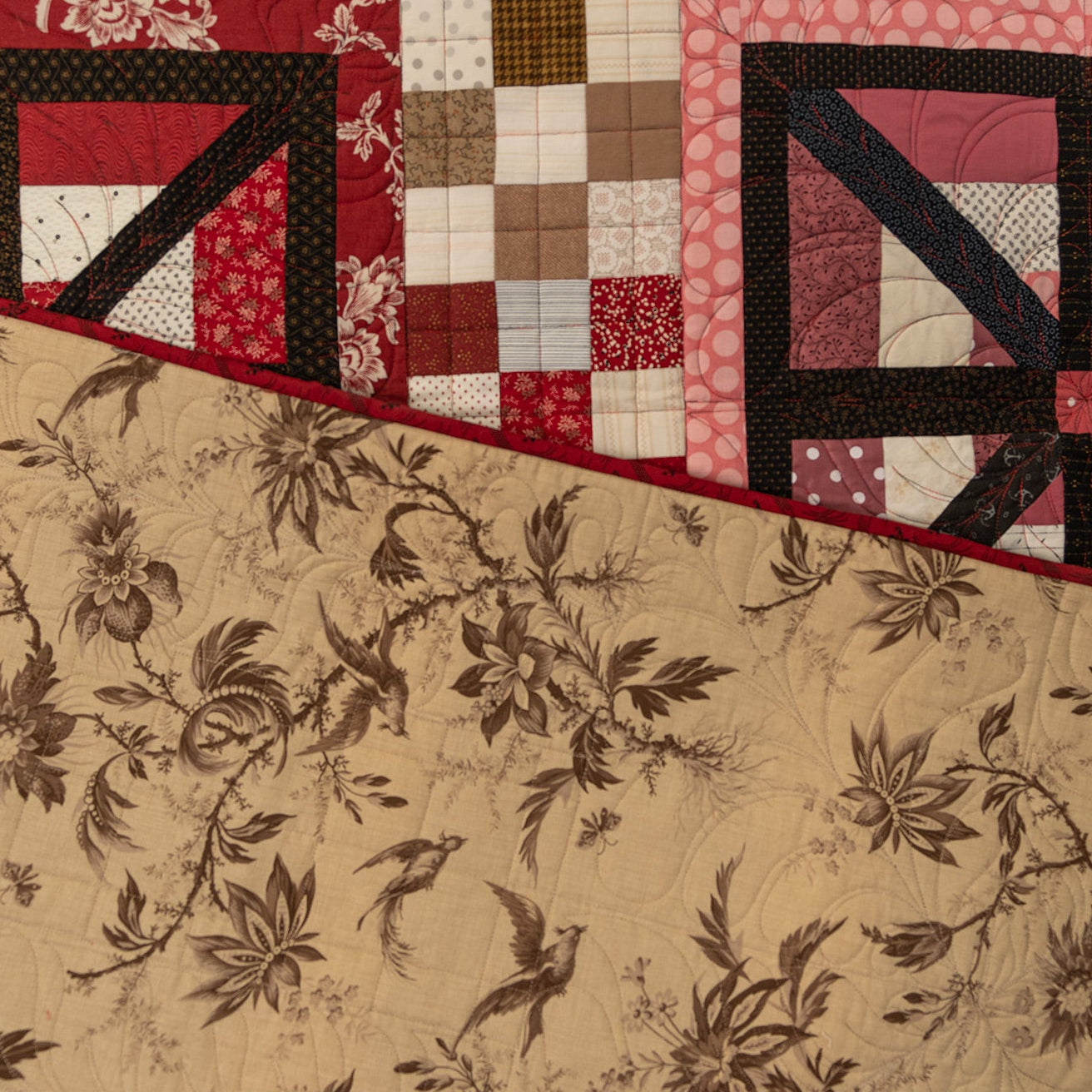A close up of a quilt, the main side shows the square prints in black, red and pink while the back shows the tan floral print. 